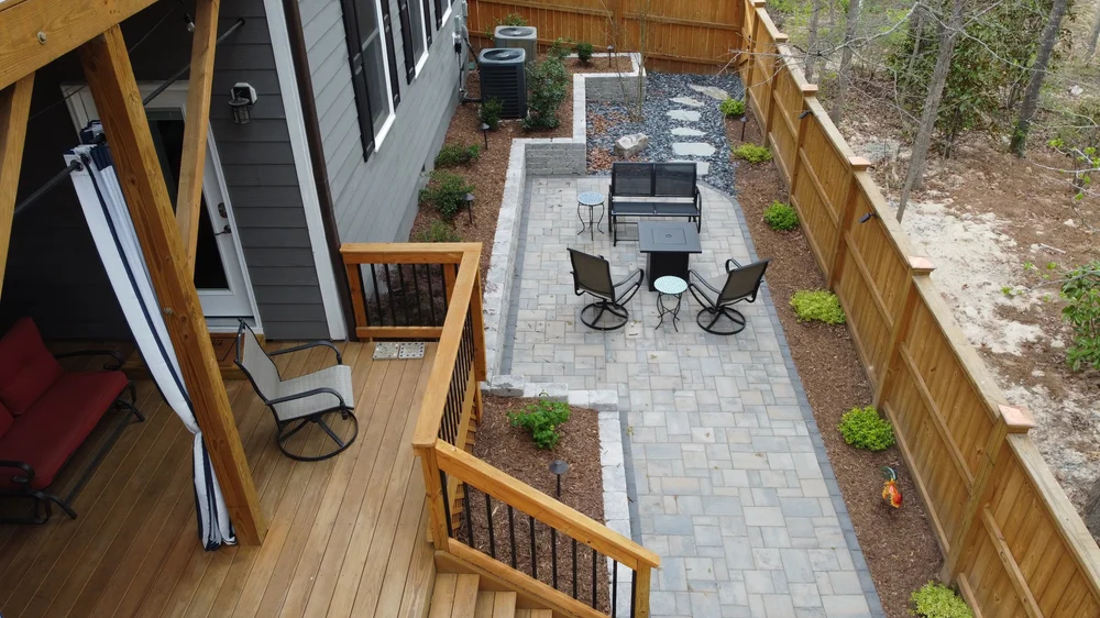 A gray and dark gray patio with a small black fireplace centered between an outdoor black aluminum bench and two swivel chairs with end tables. A large light brown stained deck with outdoor furniture and a weston stone retaining wall and wooden fence