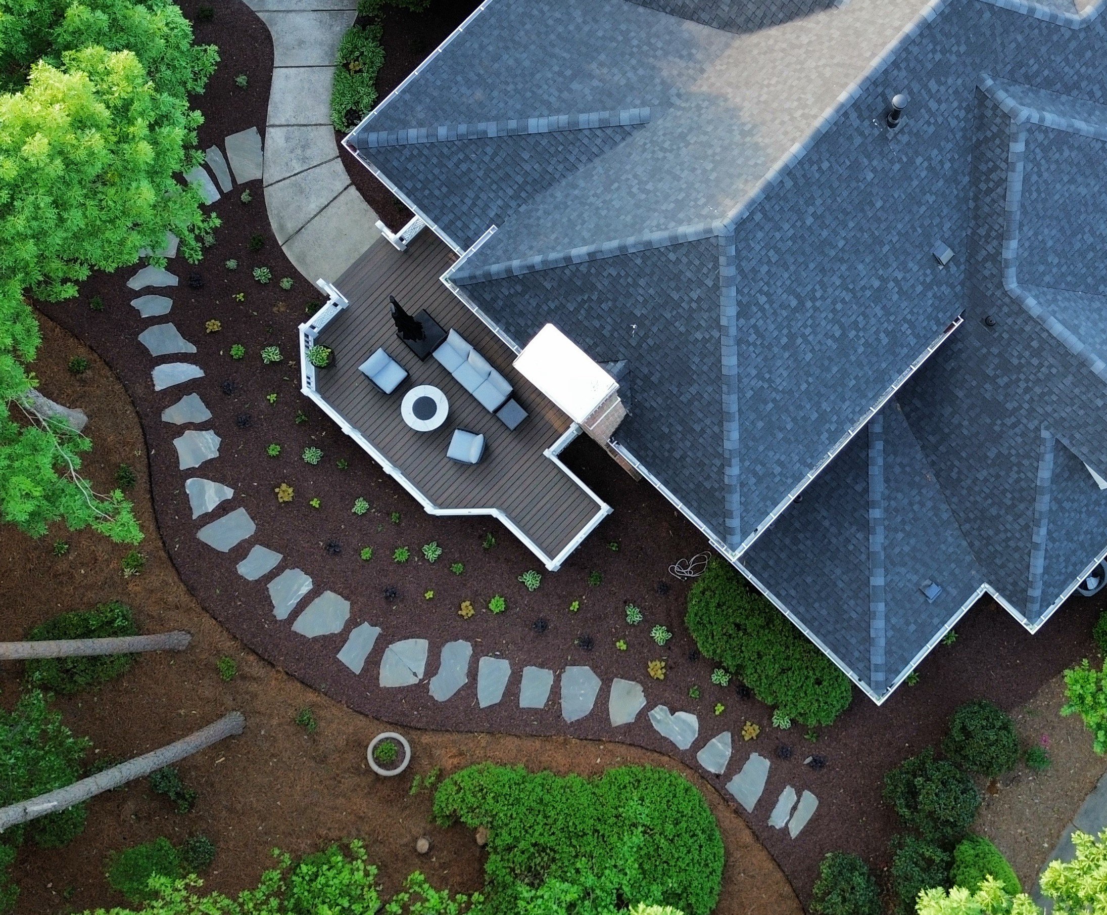 a birds eye view of a homes backyard with mulch and trees, plants of green, yellow, and purple leaves in front of a trex board deck with outdoor furniture and a clean concrete and separate flagstone walkway summer entertaining outdoor living space