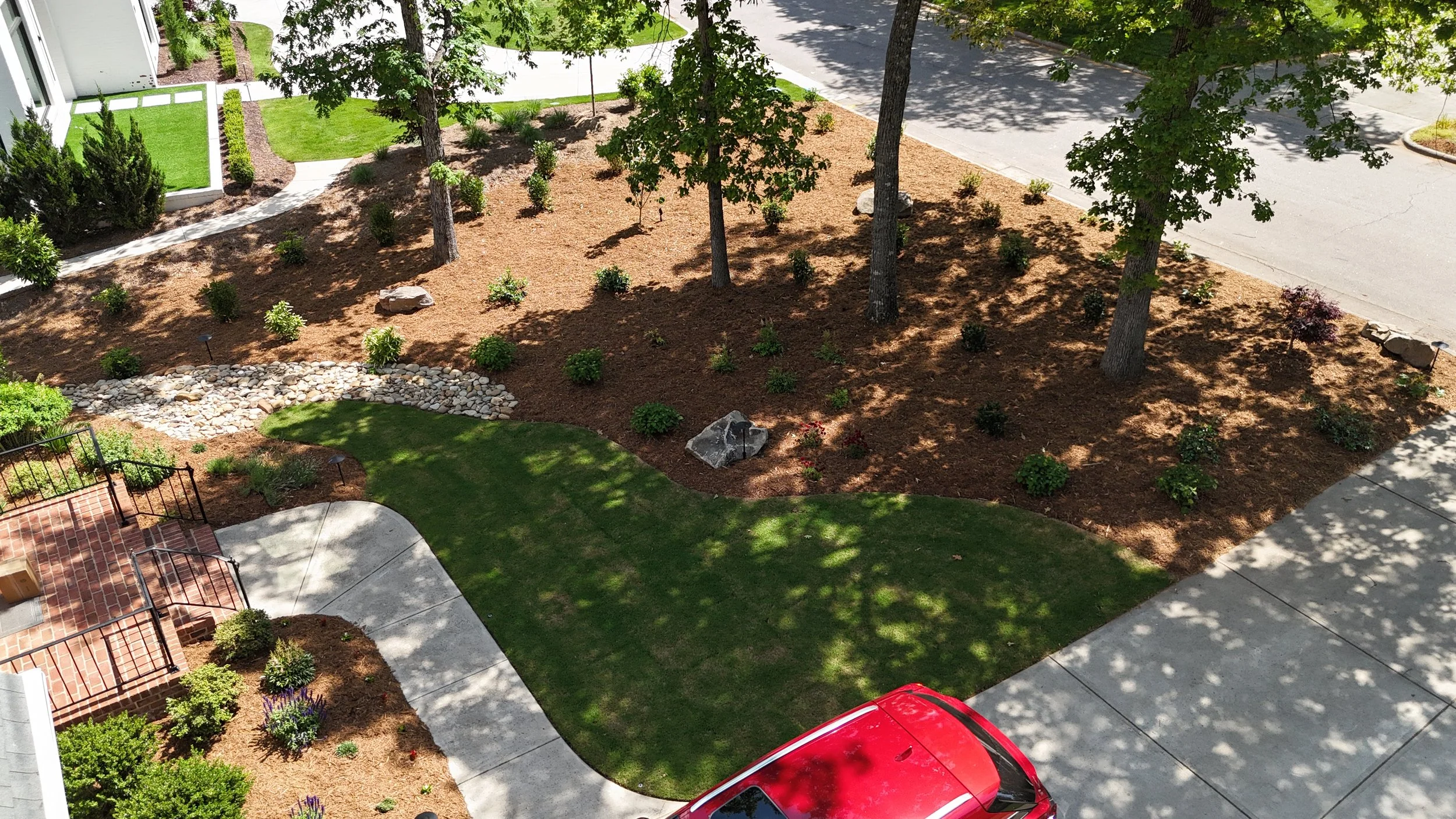 a green lawn with pine straw covered garden bed with plants and boulder stones anda dry creek bed with river rock and concrete walkway leading to a front stoop with black railings in front yard