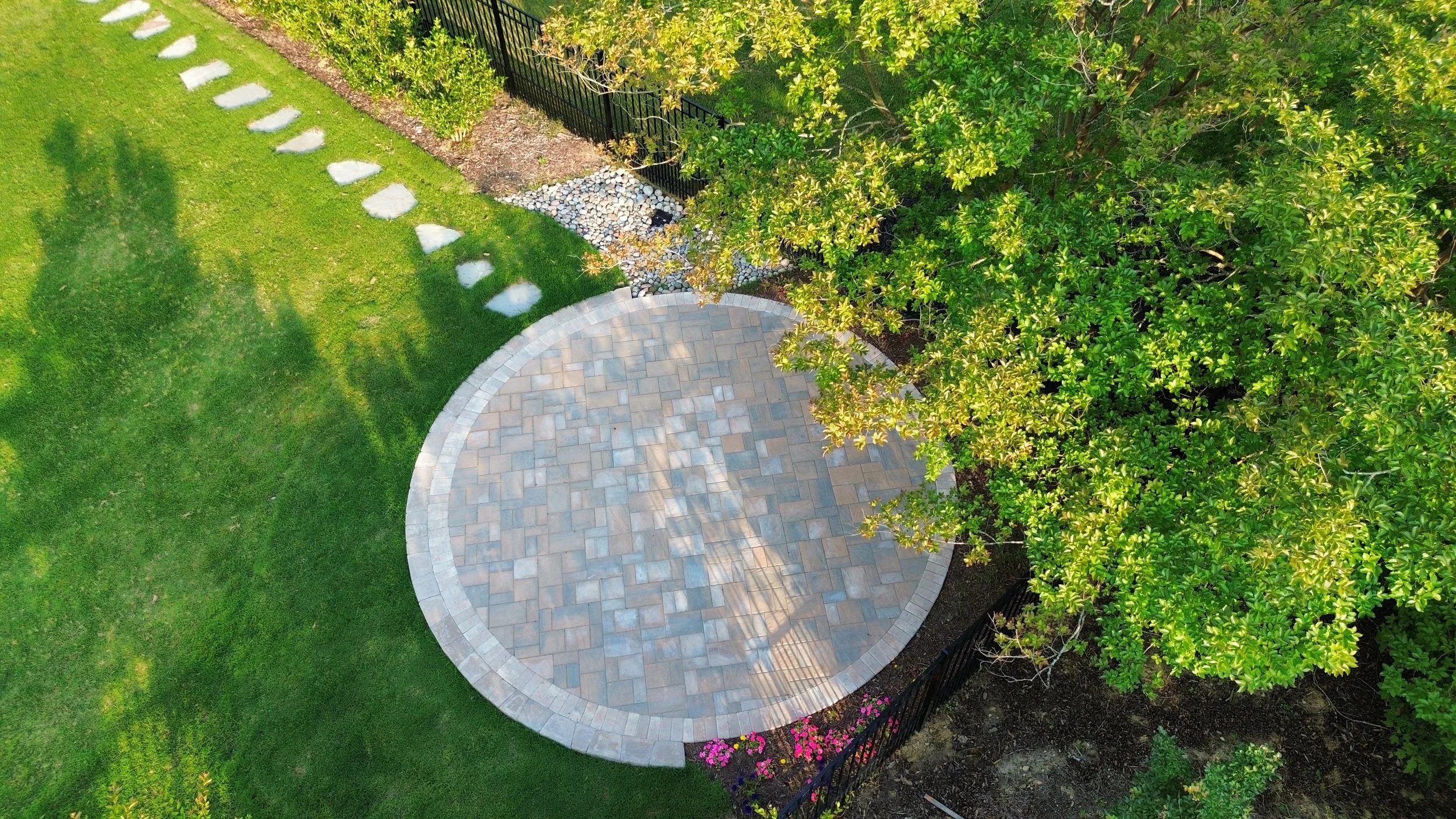 an aerial photo of a backyard with green lush sod with an embedded circular paver patio with brown and grey colorways, with a tree with yellowing leaves and small mulch area.