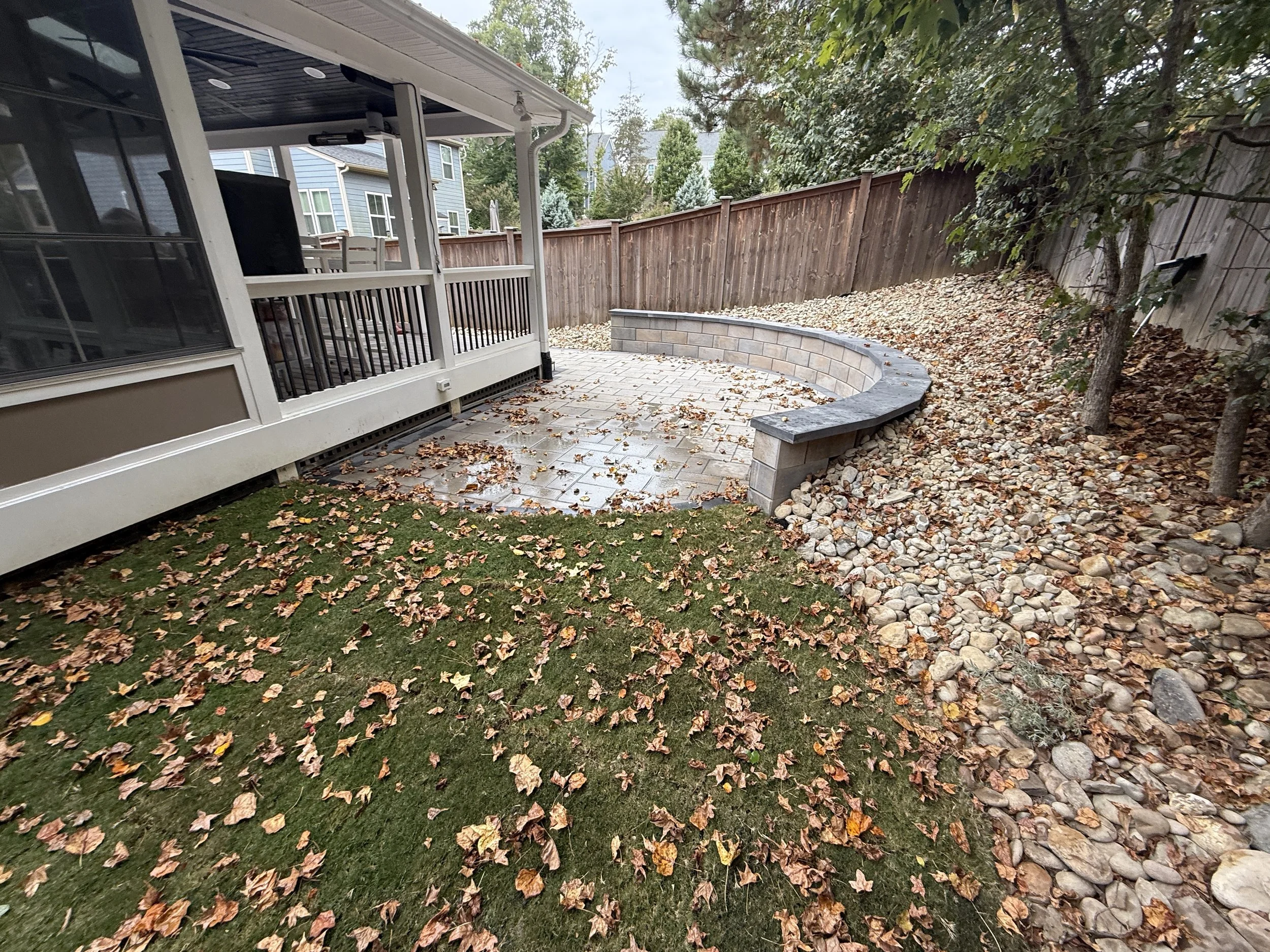 Fresh landscaped sod installation sprinkled with yellow, orange fallen leaves next to a paver patio and techo bloch g-force retaining wall retainings a dry creek bed filled with Pennsylvania river rock in the backyard of a home in apex north carolina