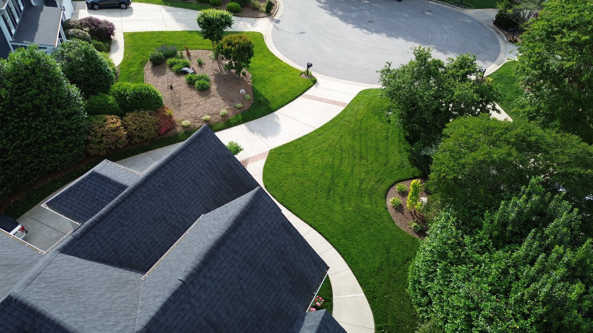 An aerial view beautiful green grass with smooth curved mulched beds on the right side and trees filling in a mature landscape and a clean driveway with brick accent bands and a roof of a house with a beautiful landscape in the front bed yard