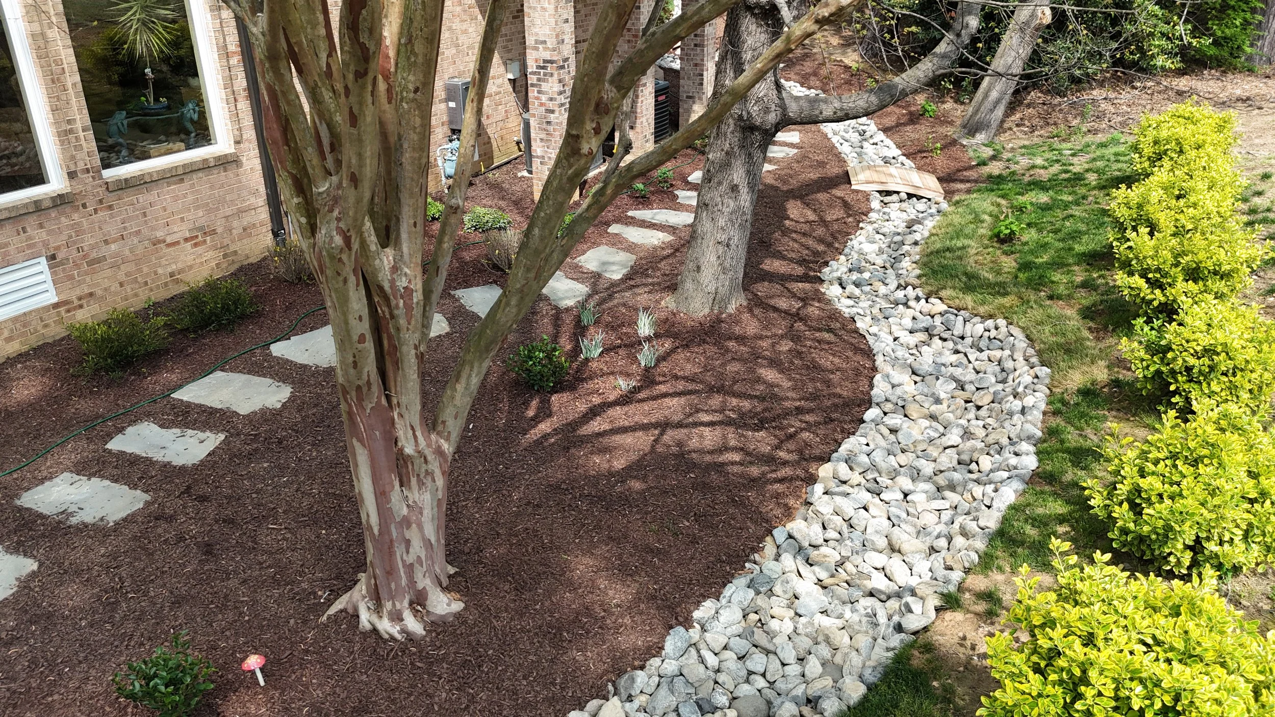 brown mulch ground cover with embedded gray irregular flagstone, trees, and a dry creek bed of 3-7 inch pensylvania river stone in a curvy shape with a wooden bridge in the side yard of a tan brick home and green fescue grass