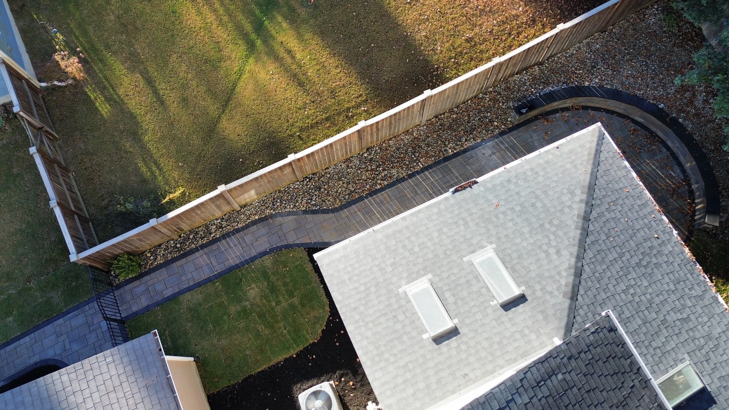 An aerial photo of a narrow side yard with a grey inlay stone paver from techo bloc with a dark ash border stone paver, green lush sod, pennsylvania river rock bed with a wooden fence and black aluminum fence and a few green plants in wake county.