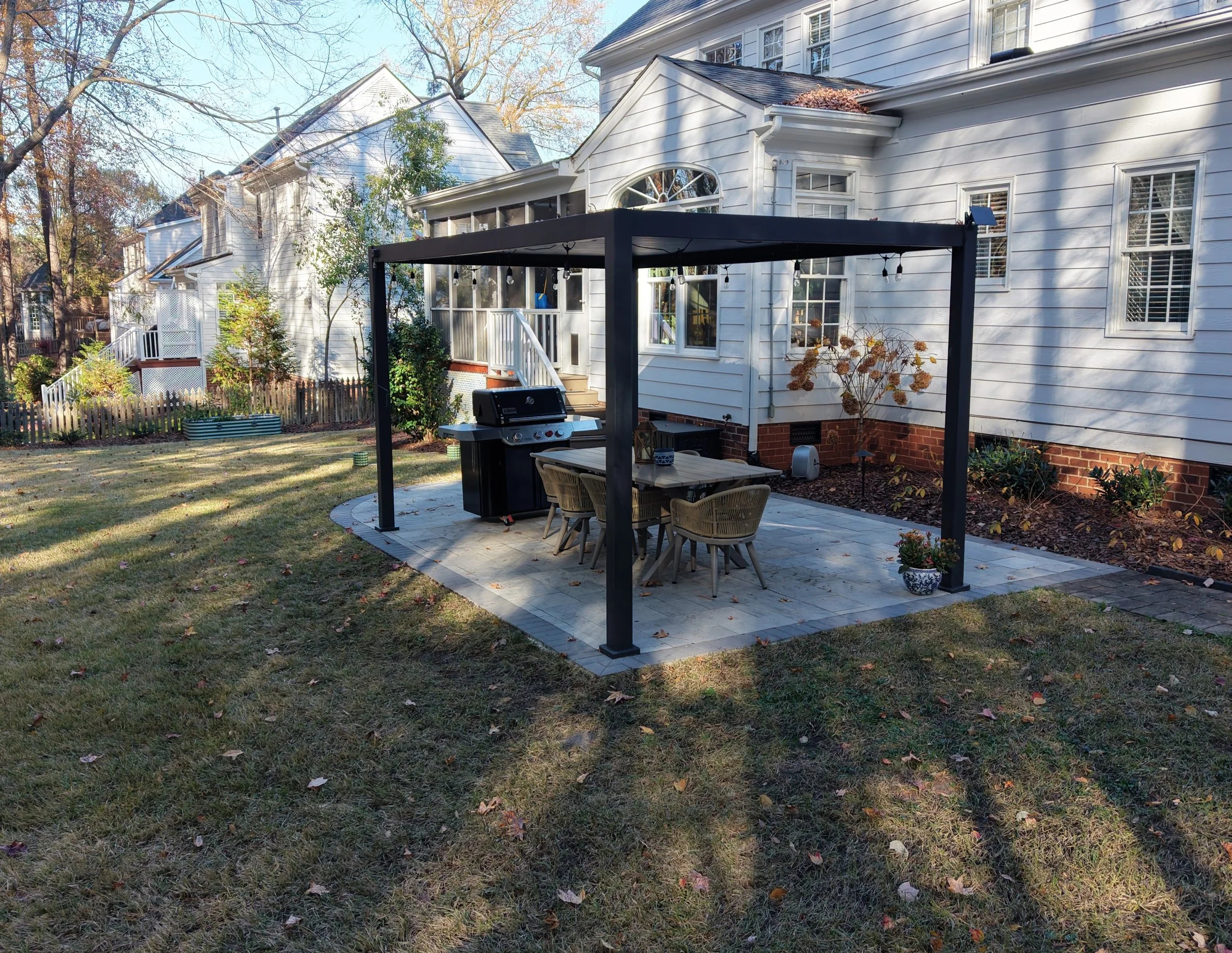A backyard with green grass sprinkled with fallen leaves with a grey paver patio covered by a black aluminum pergola, outdoor grill, outdoor dining table, and dormant plants surrounding the patio. visible along the edges of the backyard wooden fence