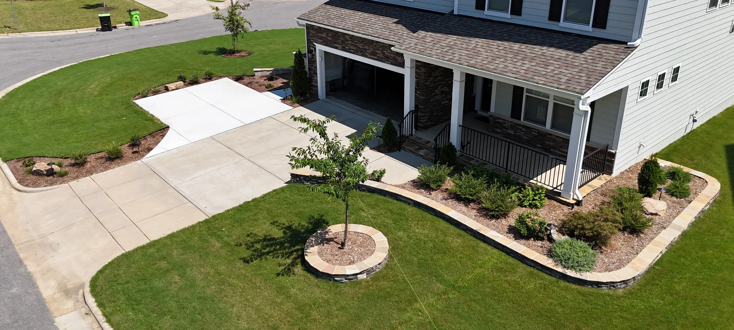 a front yard and concrete driveway with green lush grass natural mulch beds sprinkled with small evergreen plants and small boulders with veneer small retaining walls infront of a porch with metal railing and white columns and an open garage