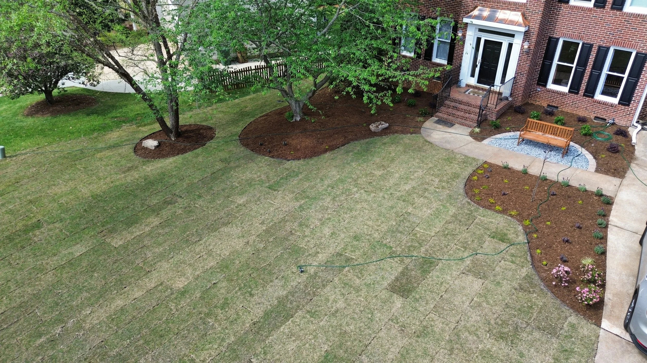 green water hoses spread across a front yard over green grass, brown mulch beds with plants, trees, and a bench over gravel in cary nc landscape