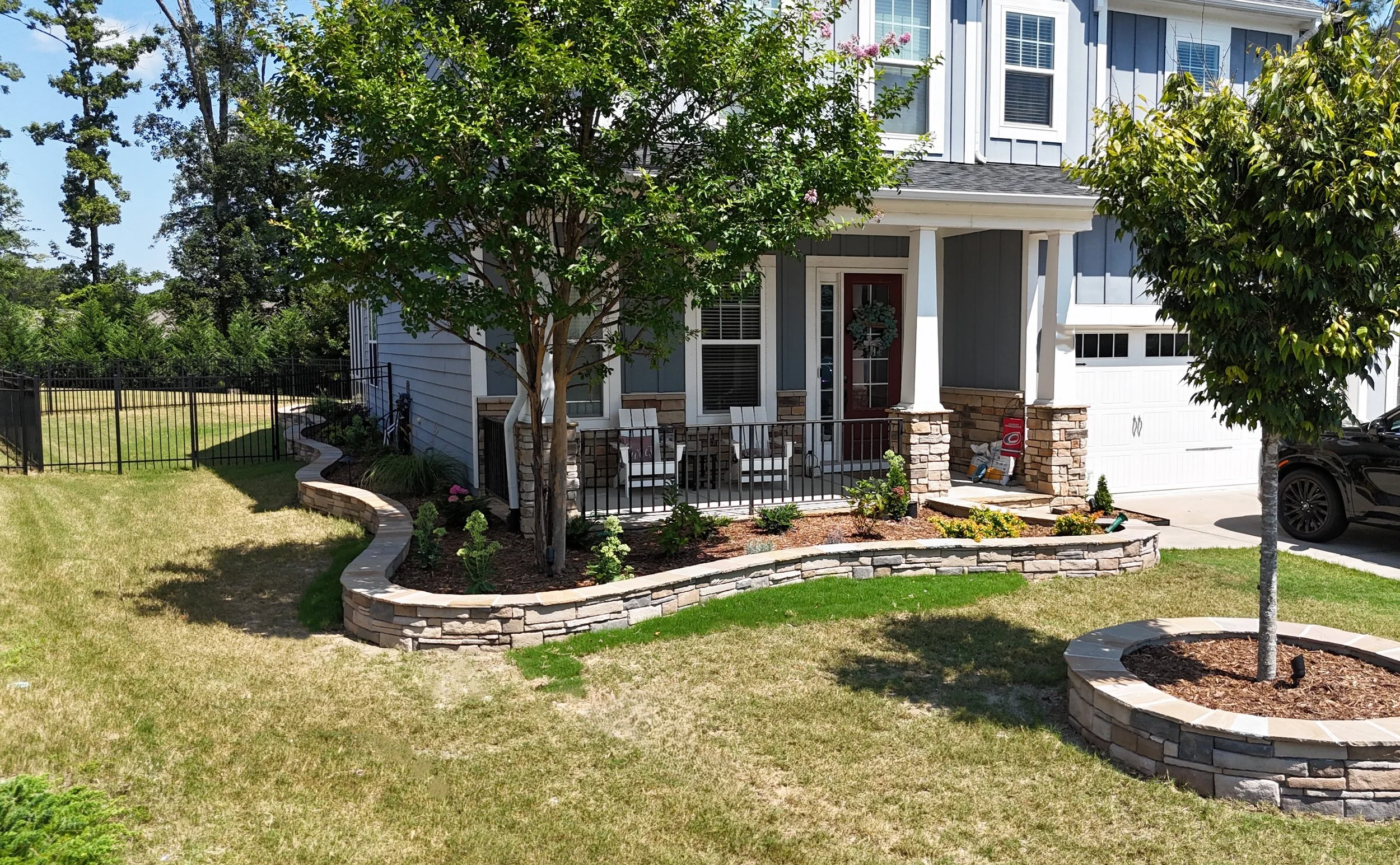 front yard and porch of a single family home, with focus on a veneer retaining wall, with a solstace cap encslosing mulch and perennial and ever green plants with blooming flowers green, yellow, pink, and blue surrounded by green sod