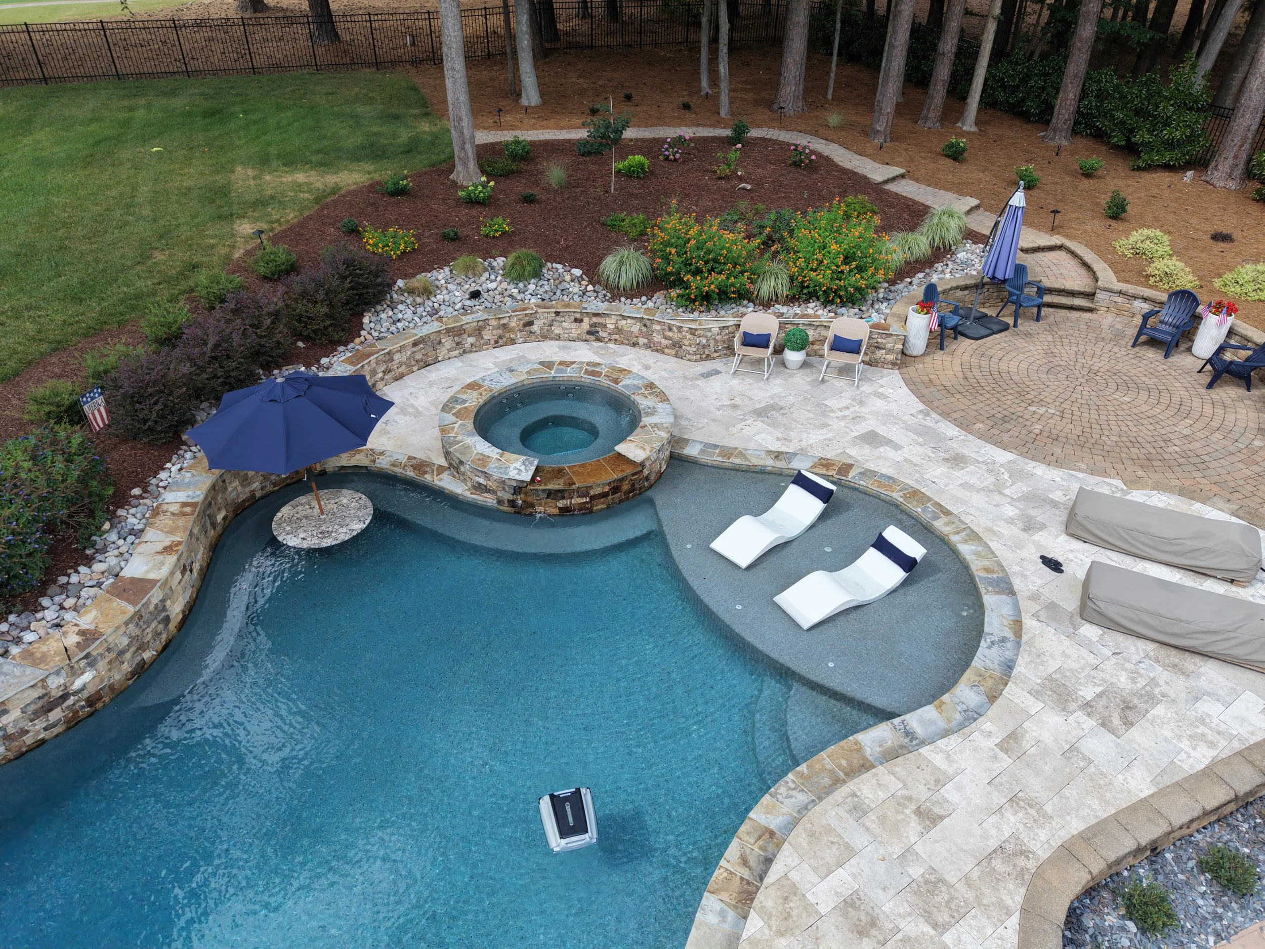 an aerial view of a curved natural stone coping pool with beautiful grey and light brown and blue tile with riverstone outlining the pool and a blue pool umbrella and white pool loungers and grass along the edge of the pool.
