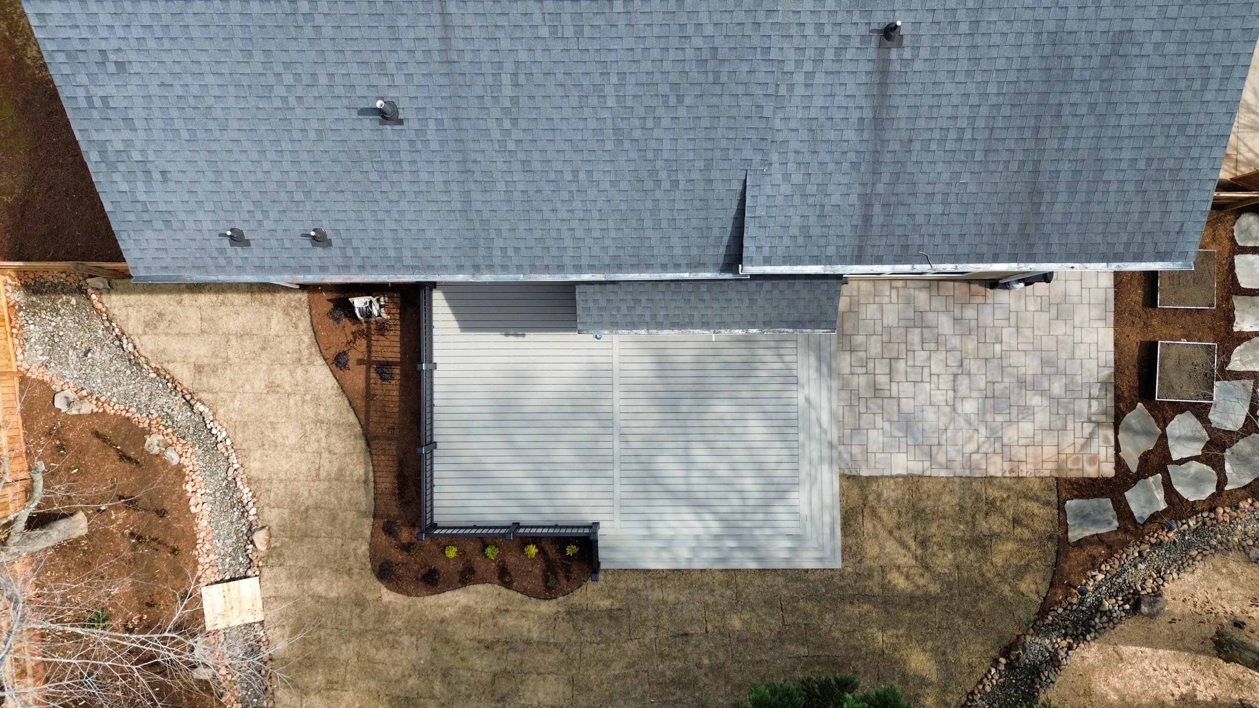 an aerial view of a small backyard with a intentionally designed deck low to the ground with a small paver patio with fresh sod for a lawn and mulch area and dry creek bed. The deck is a cool grey color using trex boards with a black aluminum railing