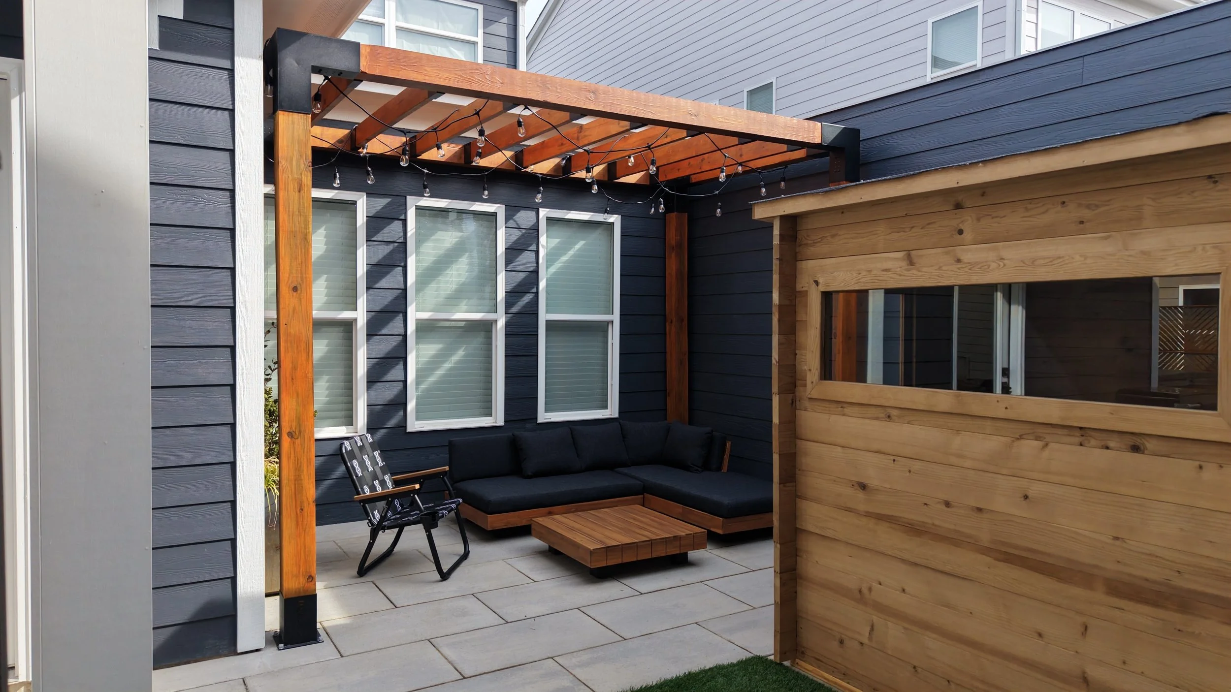 a wooden pergola stained walnut color with string lights hanging from the top of the pergola beams with toja grid corner metal posts over a paver patio with blue siding and windows and a personal sauna in a backyard in triangle area wake county nc