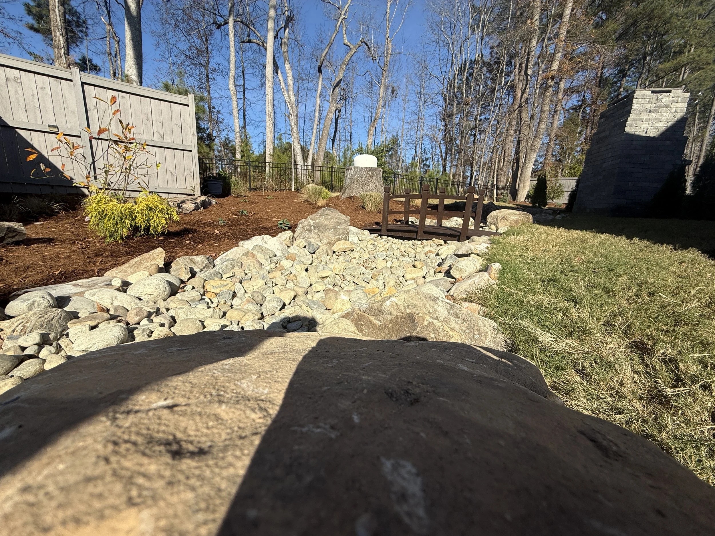 a side view of a winter water nc landscape with a boulder in a dry creek bed with a bridge connecting a mulch bed and bermuda sod in the winter time becoming dormant and dormant plants and trees
