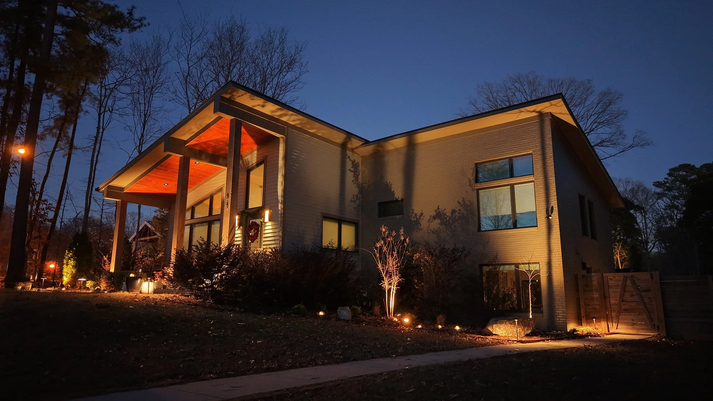 a brown and gray single family home, with landscape lights illuminating the front porch, windows, steps, yard, and front of house. There are plants and trees cascading onto the home. There is also a fence and the background sky.