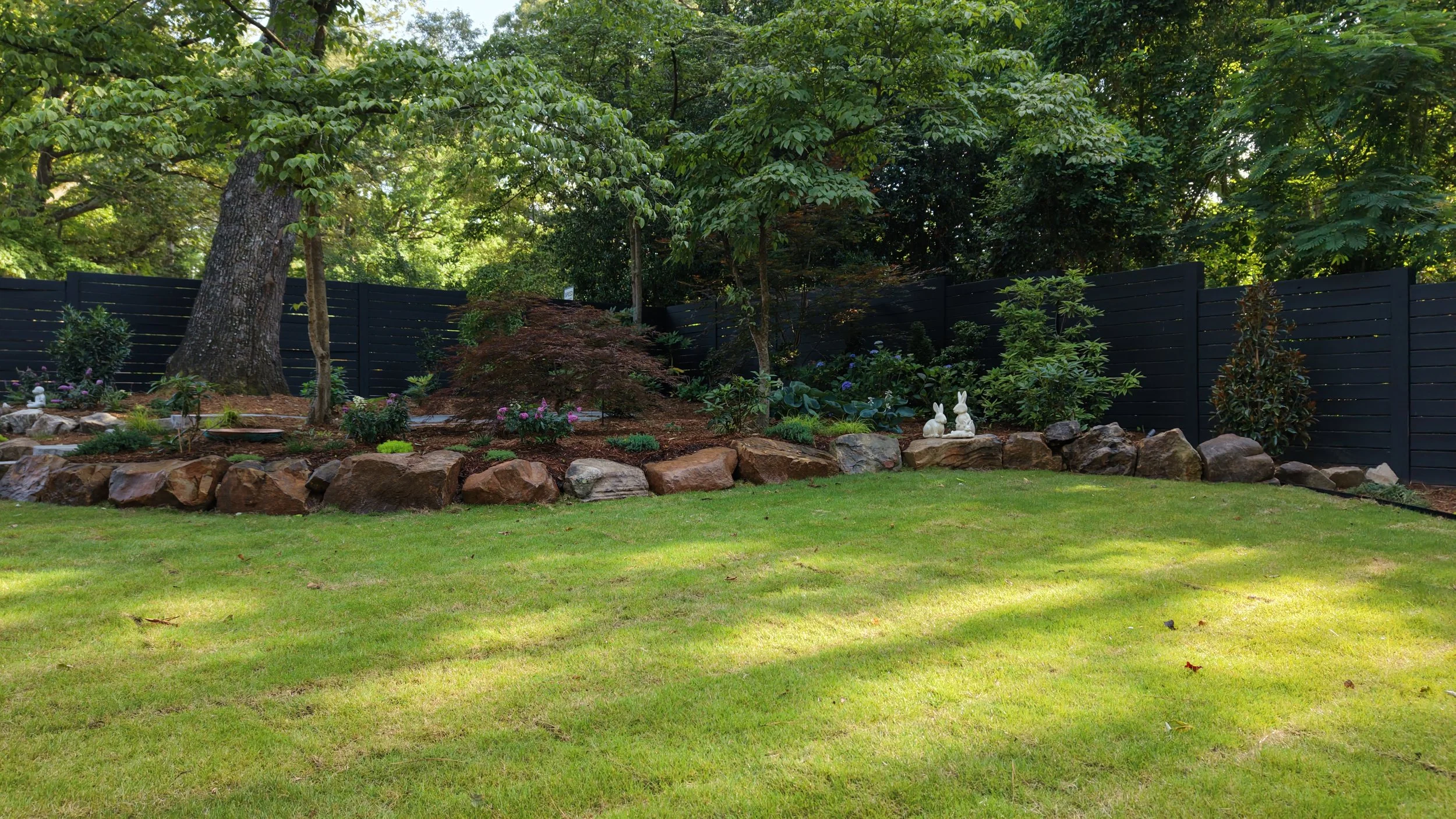 a photo of a backyard oasis with green lush warm season grass lawn with large brown boulders and variety of plants in green, purple, pink, and blue shades of color with mulch, natural boulder retaining wall, mature landscape in the triangle area nc