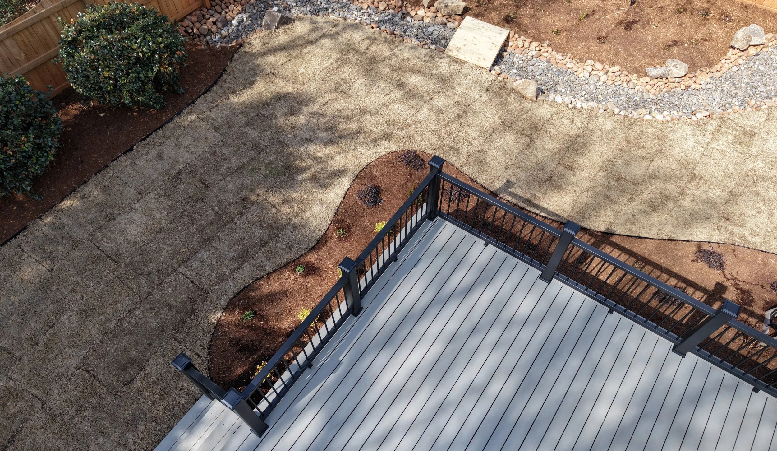 A backyard landscape from an aerial view of gray trex board deck and black aluminum railing post leading to a dormant bermuda sod lawn with a manicured landscape bed with brown mulch small shrubs in yellow, green, and purple, and black aluminum edge