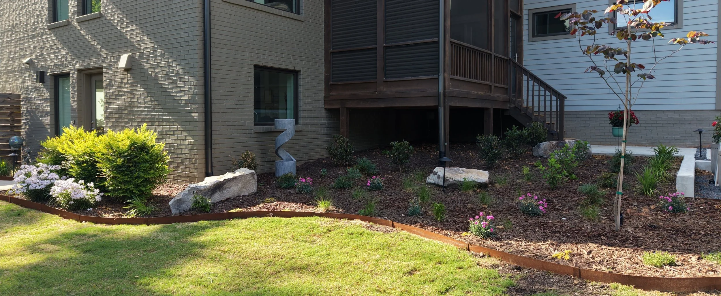 a profile photo of plantings in a natural garden bed with natural triple shred mulch and some dormant plants and boulders with a fountain against a gray brown brick contemporary house with black powder coated aluminum light fixtures in the wake nc