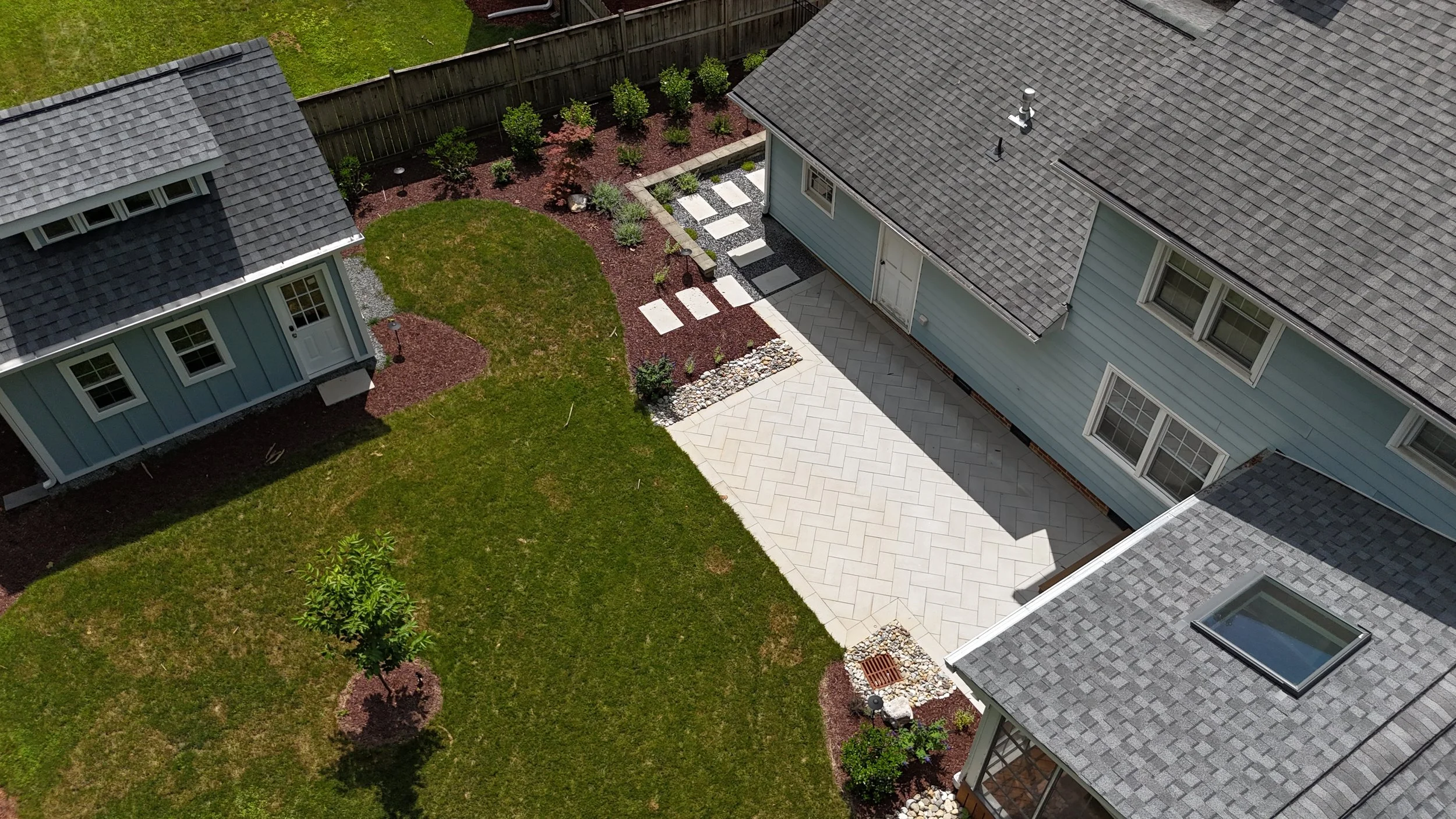 an aerial view of a backyard living space with beautiful landscaping including a off white paver stone patio in a herringbone pattern with river rock along the sides and a drainage grate embedded in green bermuda grass and dyed walnut brown mulch