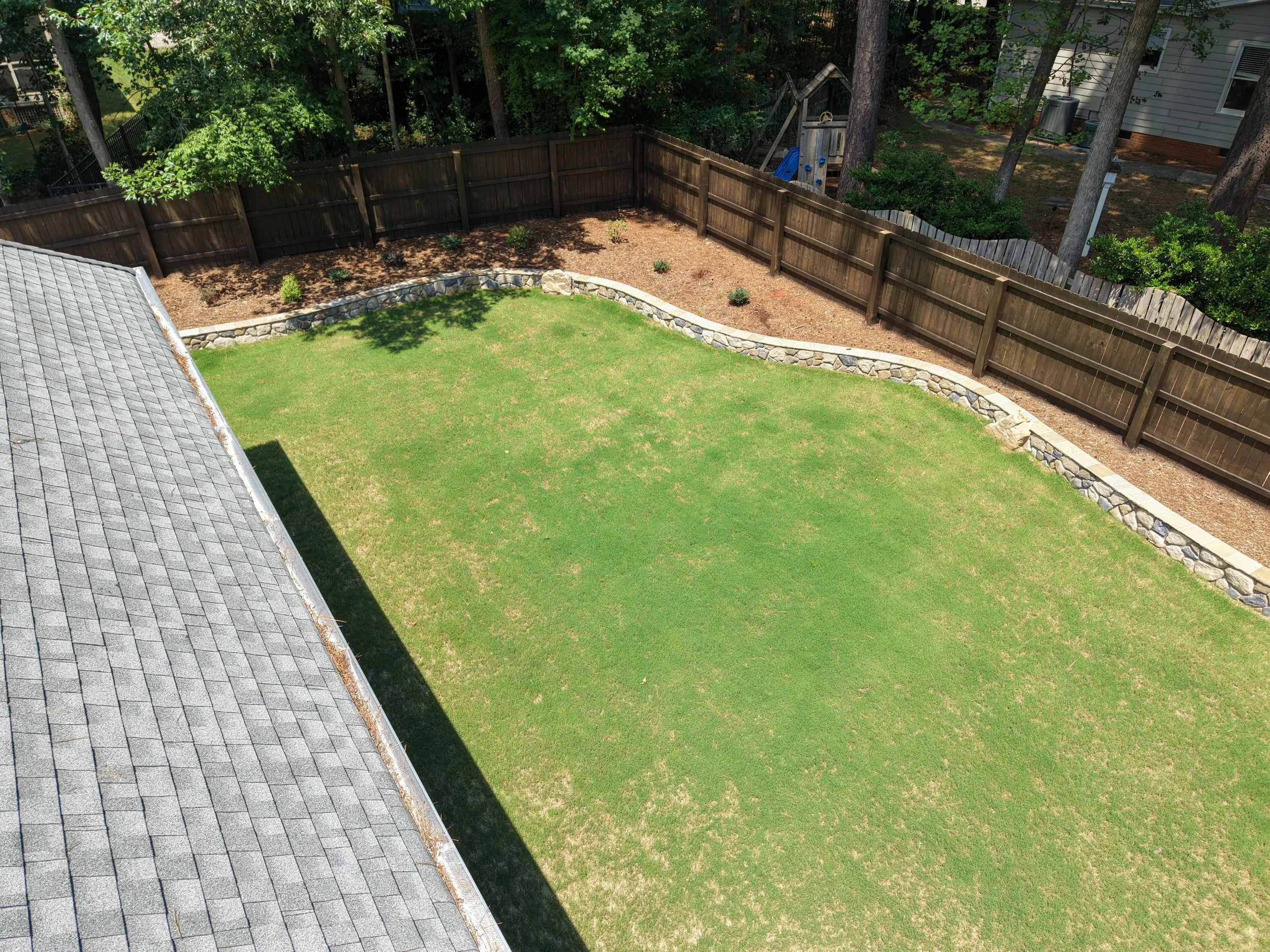 an aerial view of a backyard with green bermuda sod with patches of yellowing and browning color due to dry conditions  with a small veneer retaining wall with boulders and  plants and natural triple shred mulch in a wake county property landscaping