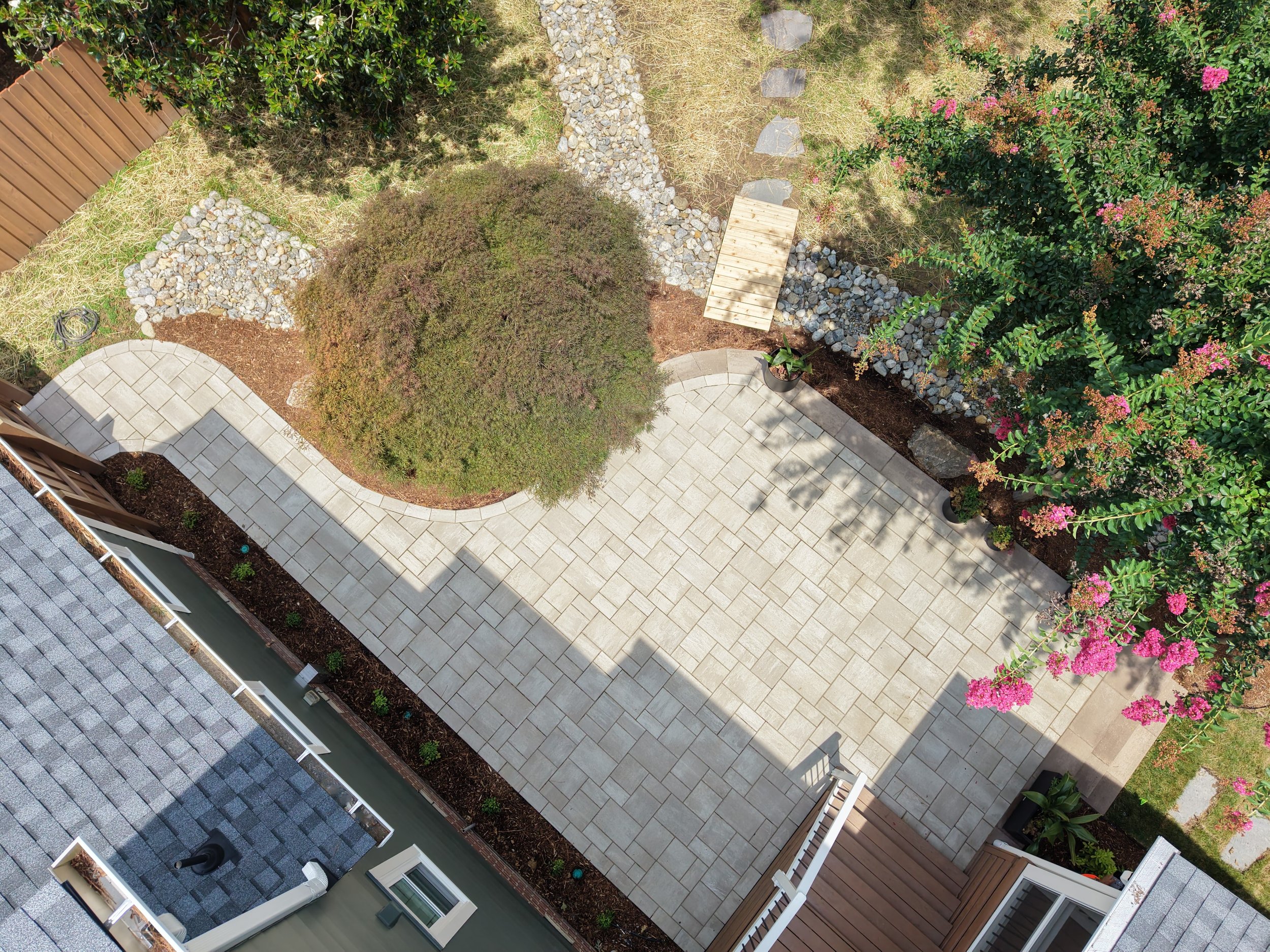 A paver patio in a backyard with natural triple shred mulch beds with green plants, a mature japanese maple plant, boulders, a pink crape myrtle, a cedar bridge and delaware river stone dry creek bed