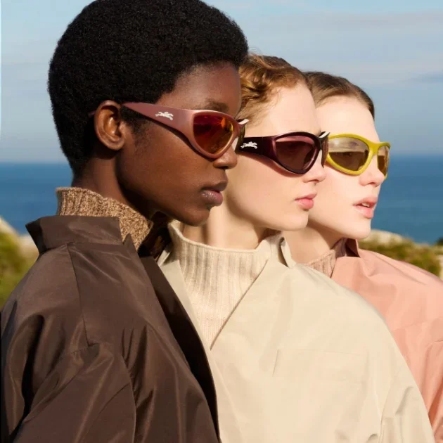 Three women standing outdoors, wearing sunglasses, with the ocean and sky in the background.