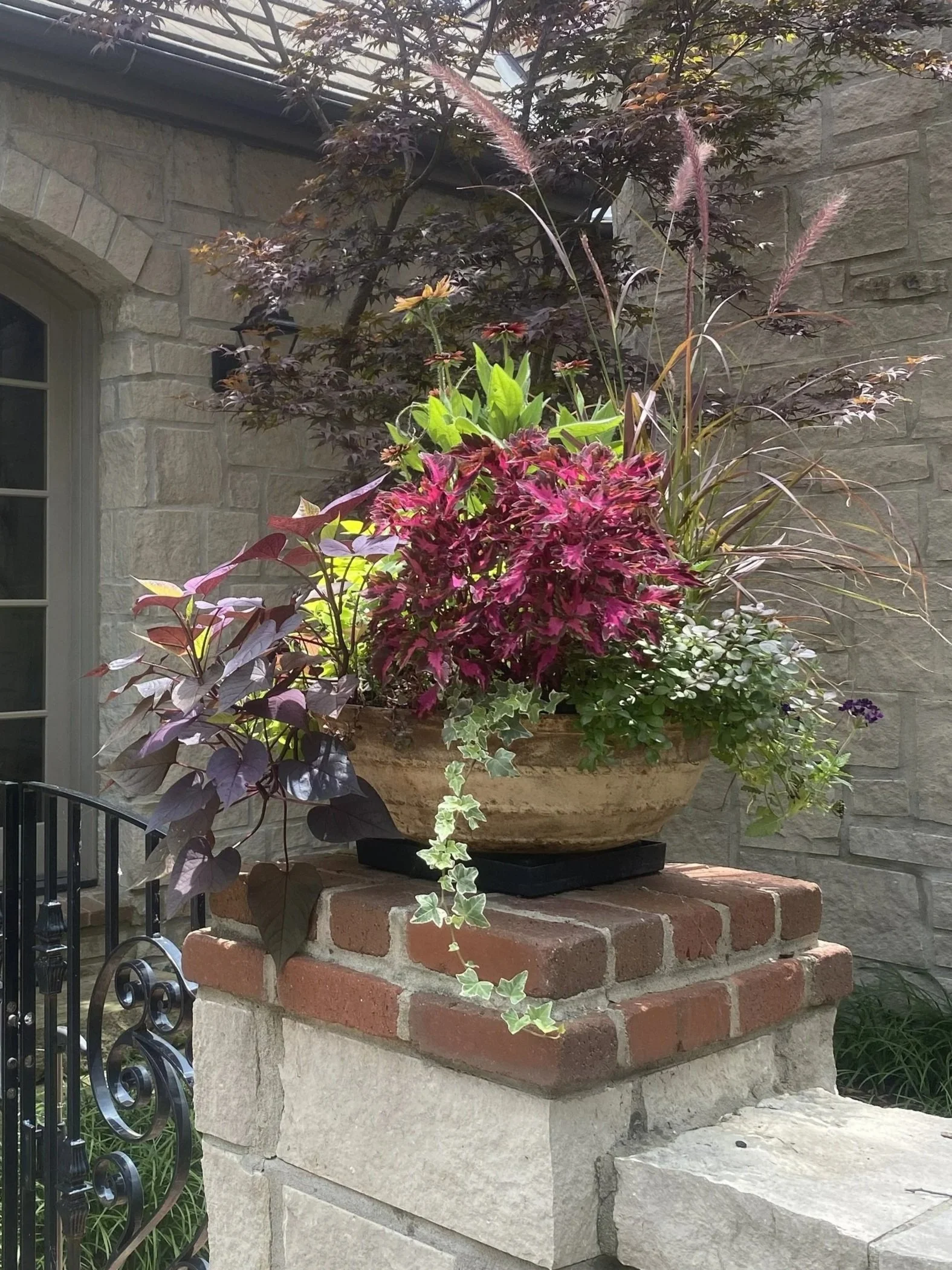A decorative flower arrangement in a large beige ceramic pot with various green, purple, and pink plants, sitting on a brick and stone pedestal outside a stone house.