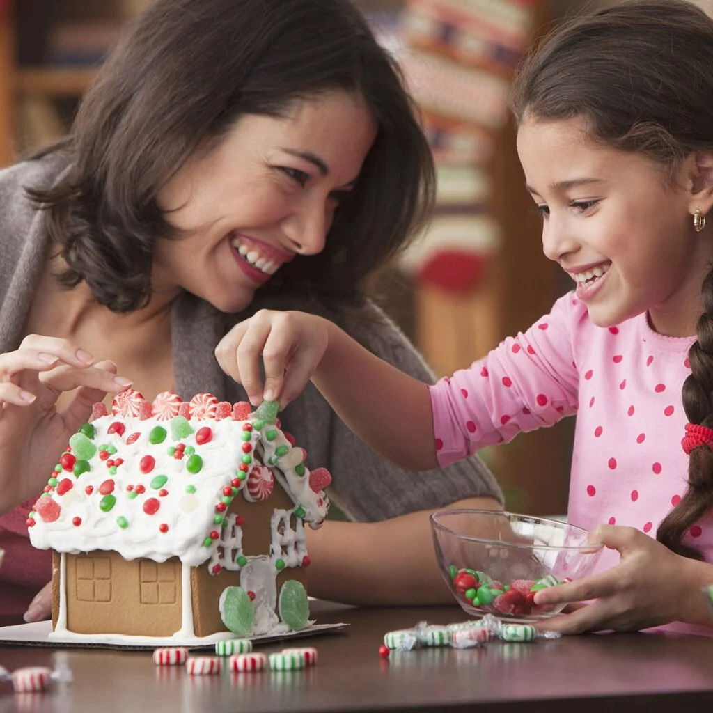 A woman and a girl decorating a gingerbread house with Christmas candies, smiling and enjoying the activity.