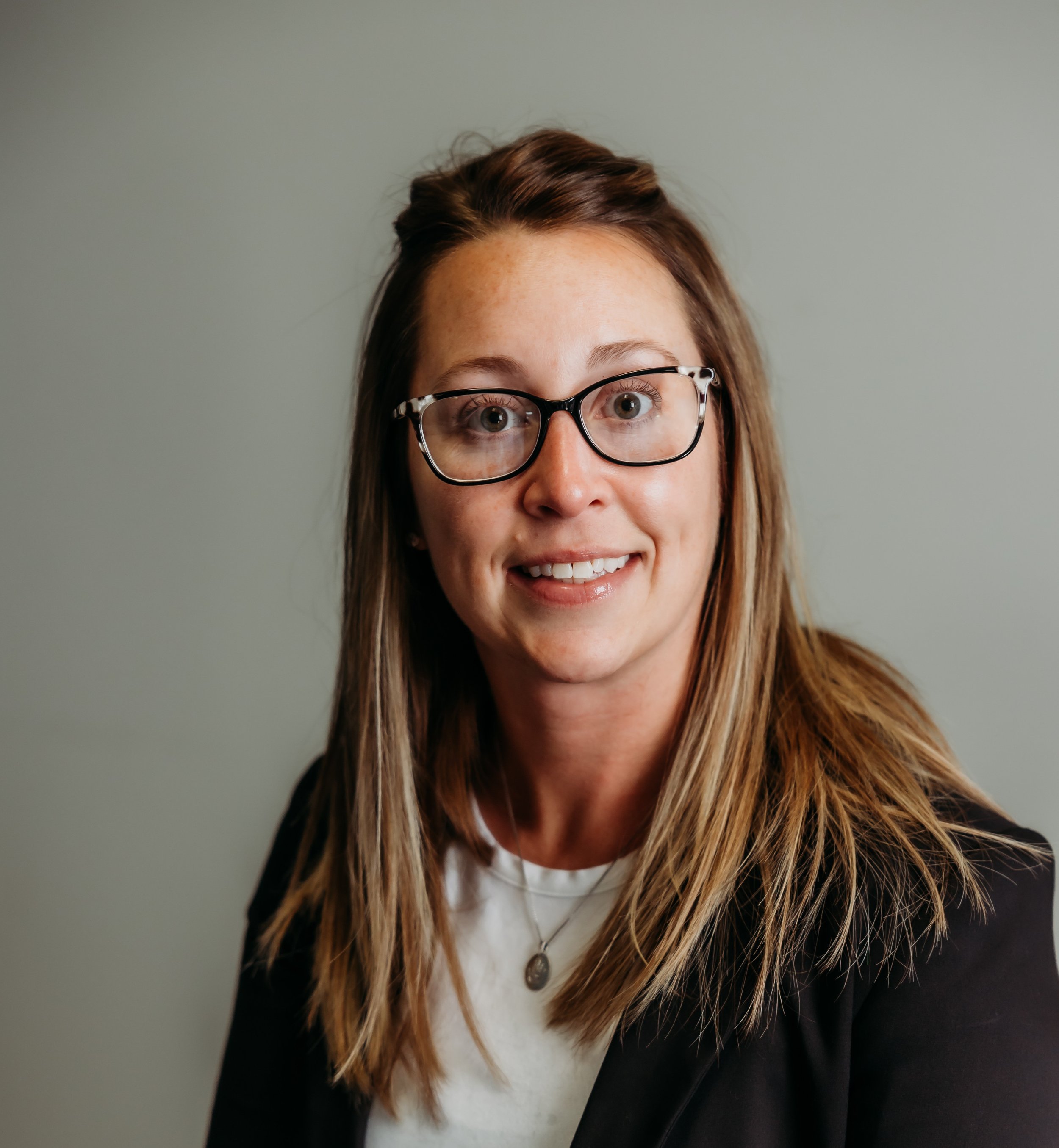 portrait of a woman with long straight hair, wearing glasses, a white top, and a dark blazer, smiling at the camera against a plain background.