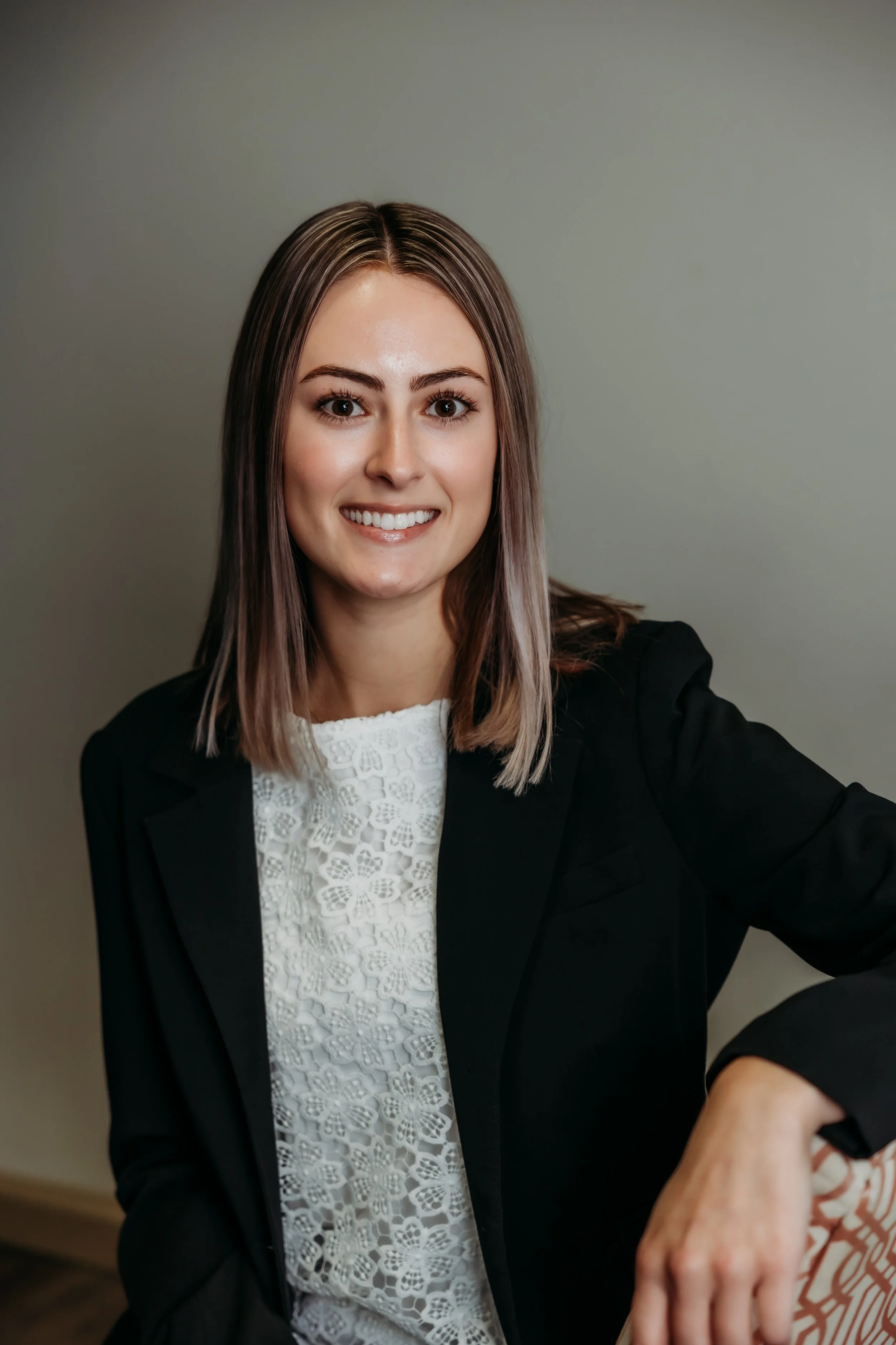 A woman with long straight hair, smiling, wearing a white top and a dark blazer, sitting on a striped chair against a plain backdrop.