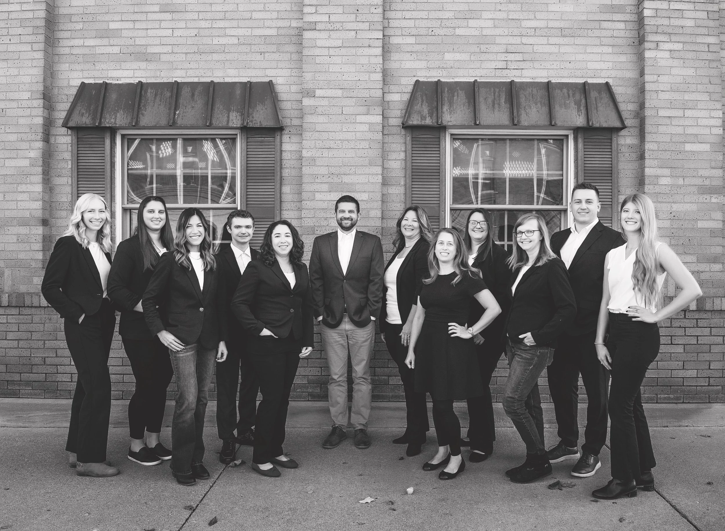 Group of fifteen people standing outdoors in front of a brick building with two windows, dressed in business attire, smiling at the camera.