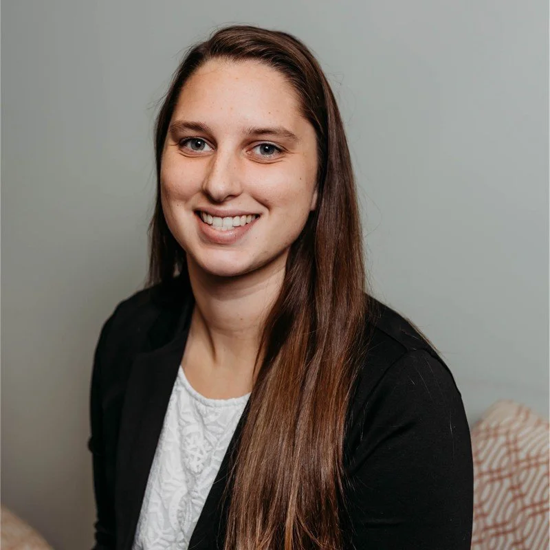 portrait of a smiling woman with long hair, wearing a light top and a dark jacket, standing against a plain background.