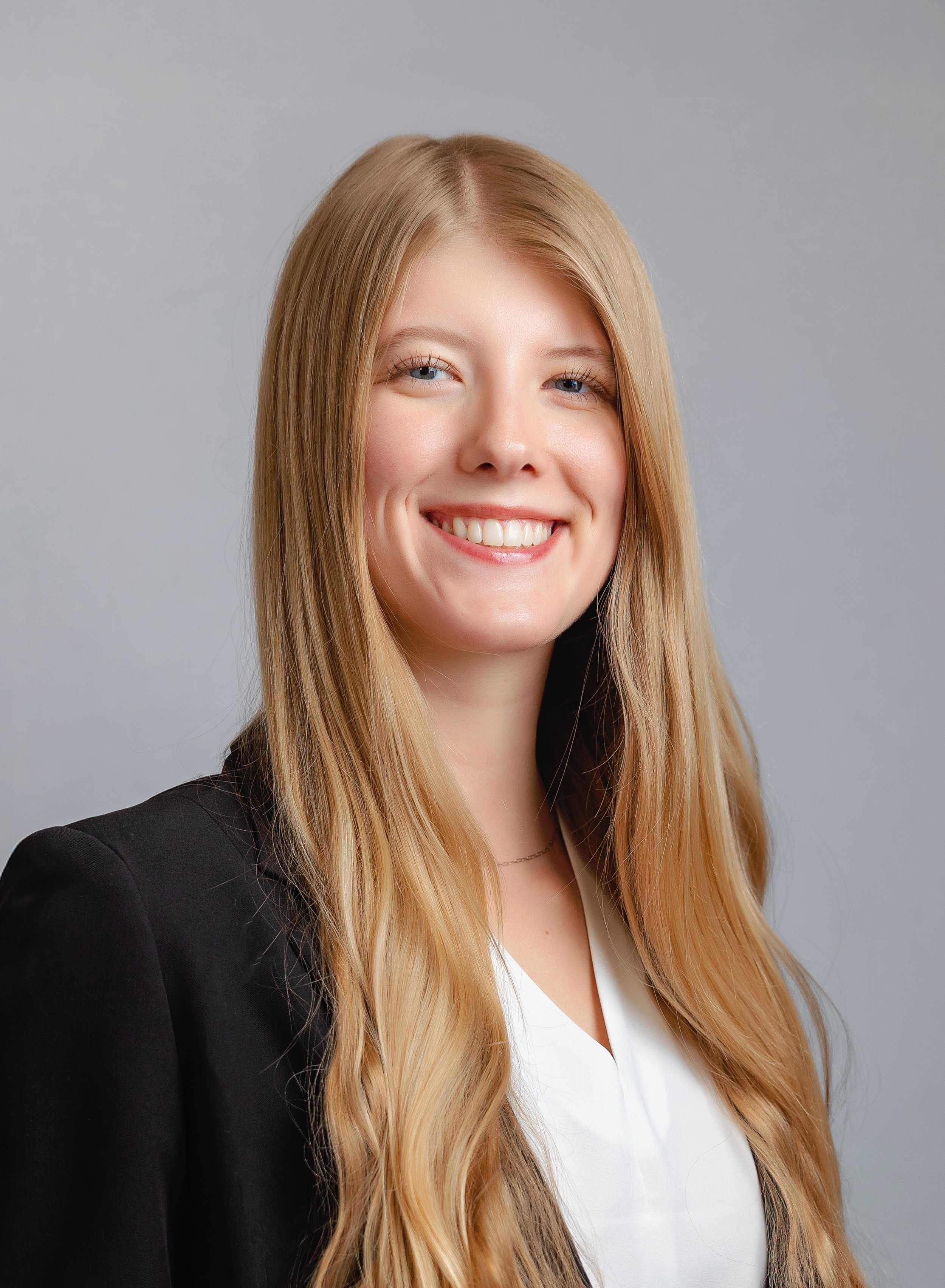 portrait of a young woman with long hair, smiling, wearing a blazer and a white top, seated with her arm resting on a patterned surface.