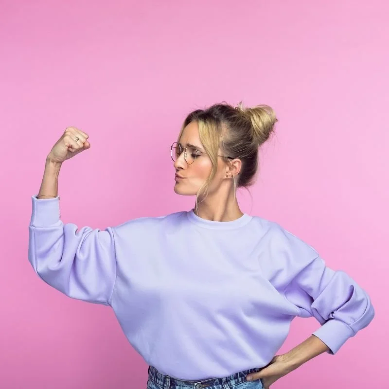 woman pumping her bicep wearing lilac colored sweatshirt in front of a easter egg pink background