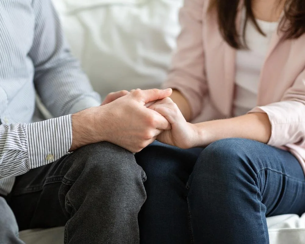 Two people holding hands, sitting close together, with their hands clasped and resting on their laps.