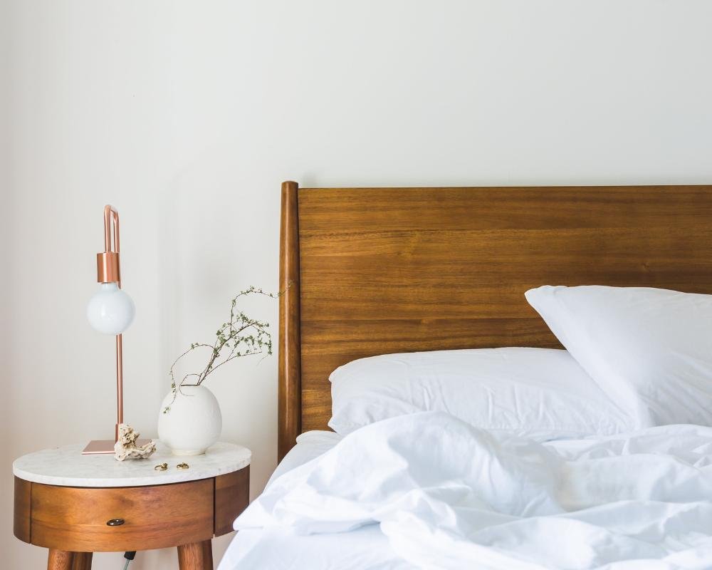 A neatly made bed with white linens, a wooden headboard, and a nightstand with a modern lamp, a small white vase with a plant, and jewelry, in a minimalist bedroom.