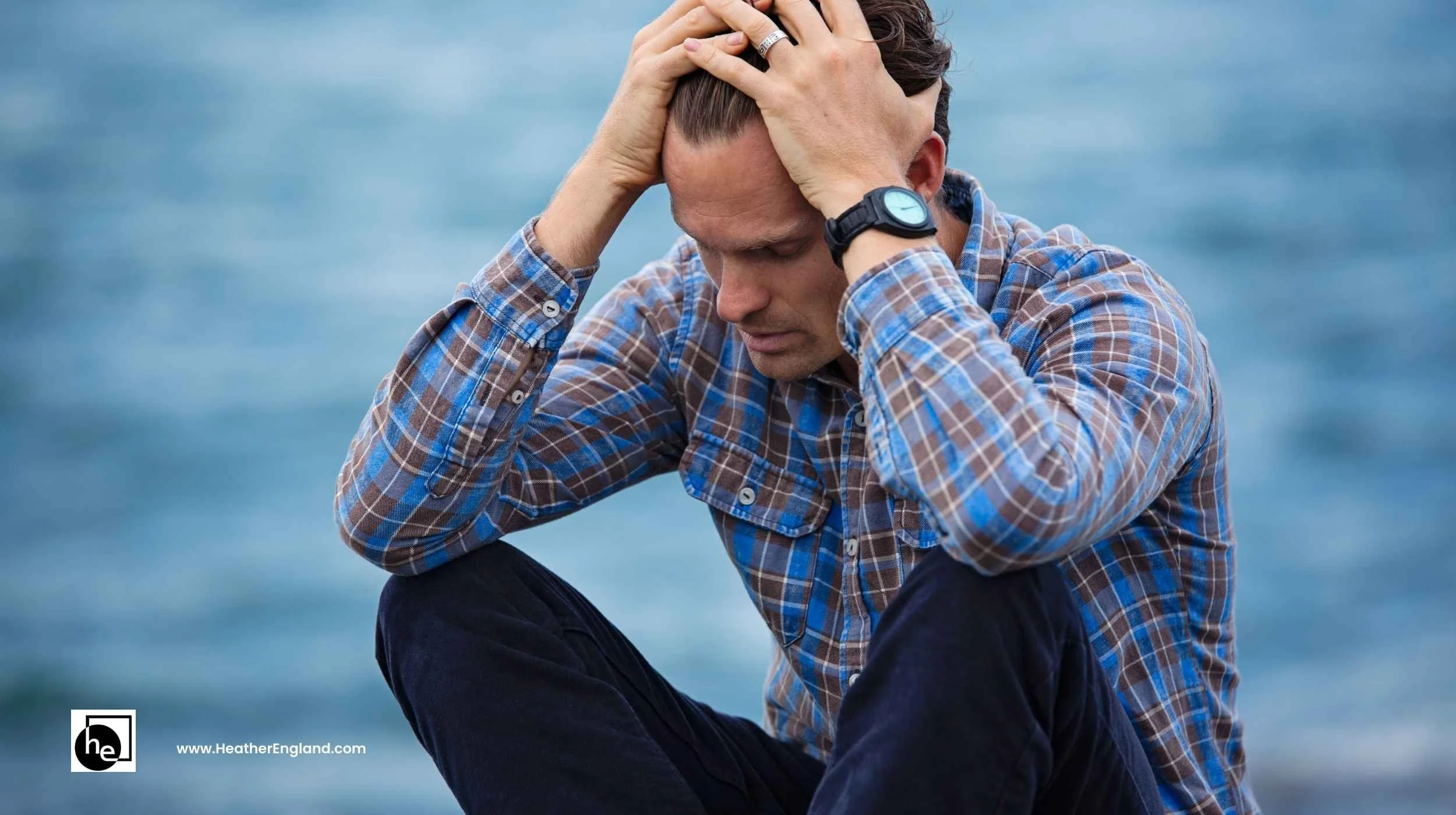 man in plaid shirt by lake holding his head and looking anxious