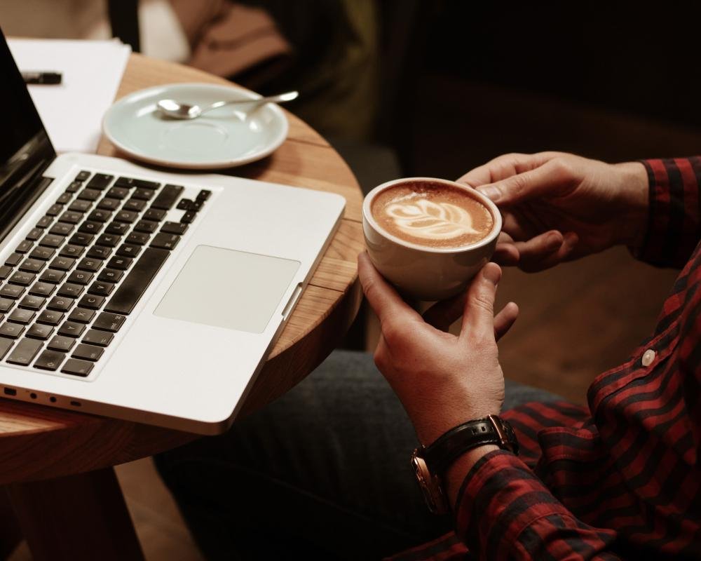 Person in a red and black striped shirt holding a cup of coffee with latte art, sitting at a wooden table with a laptop and a plate with a spoon and a fork.