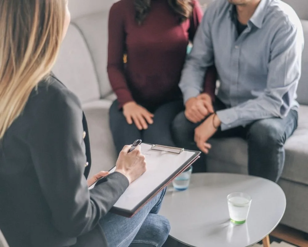 A therapist taking notes during a counseling session with a couple sitting on a sofa.
