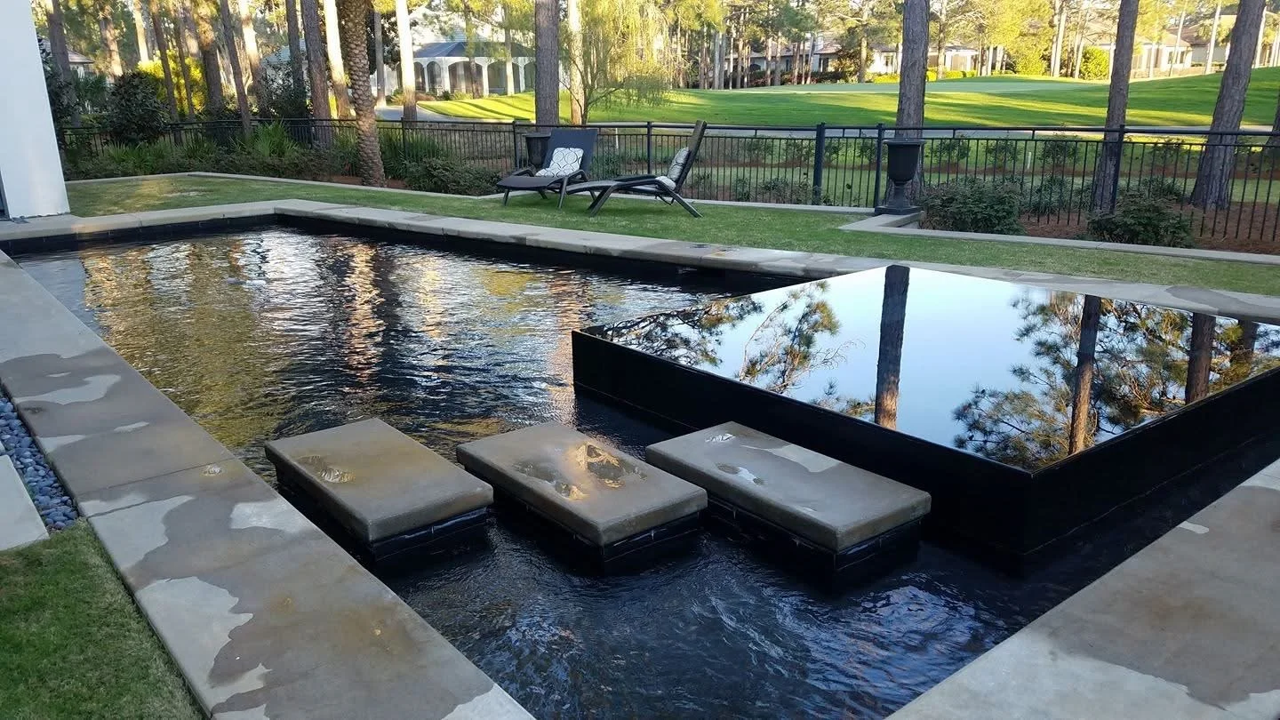 Outdoor rectangular water feature with stone steps leading into the water, surrounded by a paved border, with lawn and trees in the background, and a black fence.