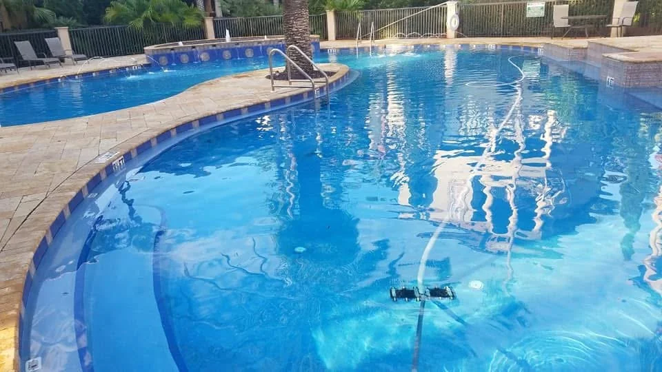 Clean swimming pool with blue water, pool cleaning equipment submerged, surrounded by a deck with lounge chairs, fencing, and trees in the background.