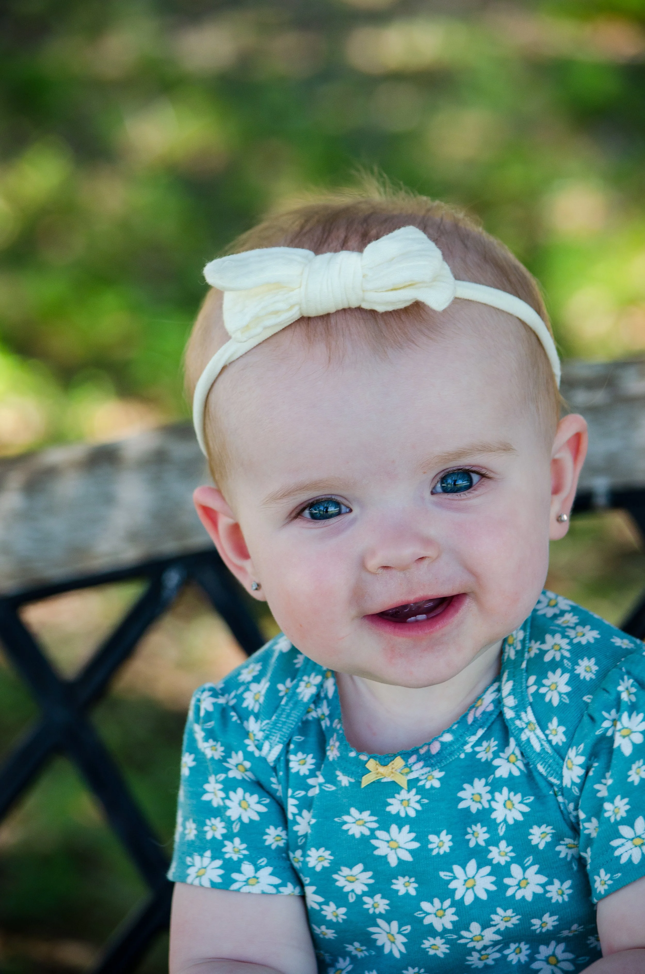 A smiling baby girl with blue eyes, wearing a blue floral dress and a white headband with a bow, outdoors on a bench with green foliage in the background.
