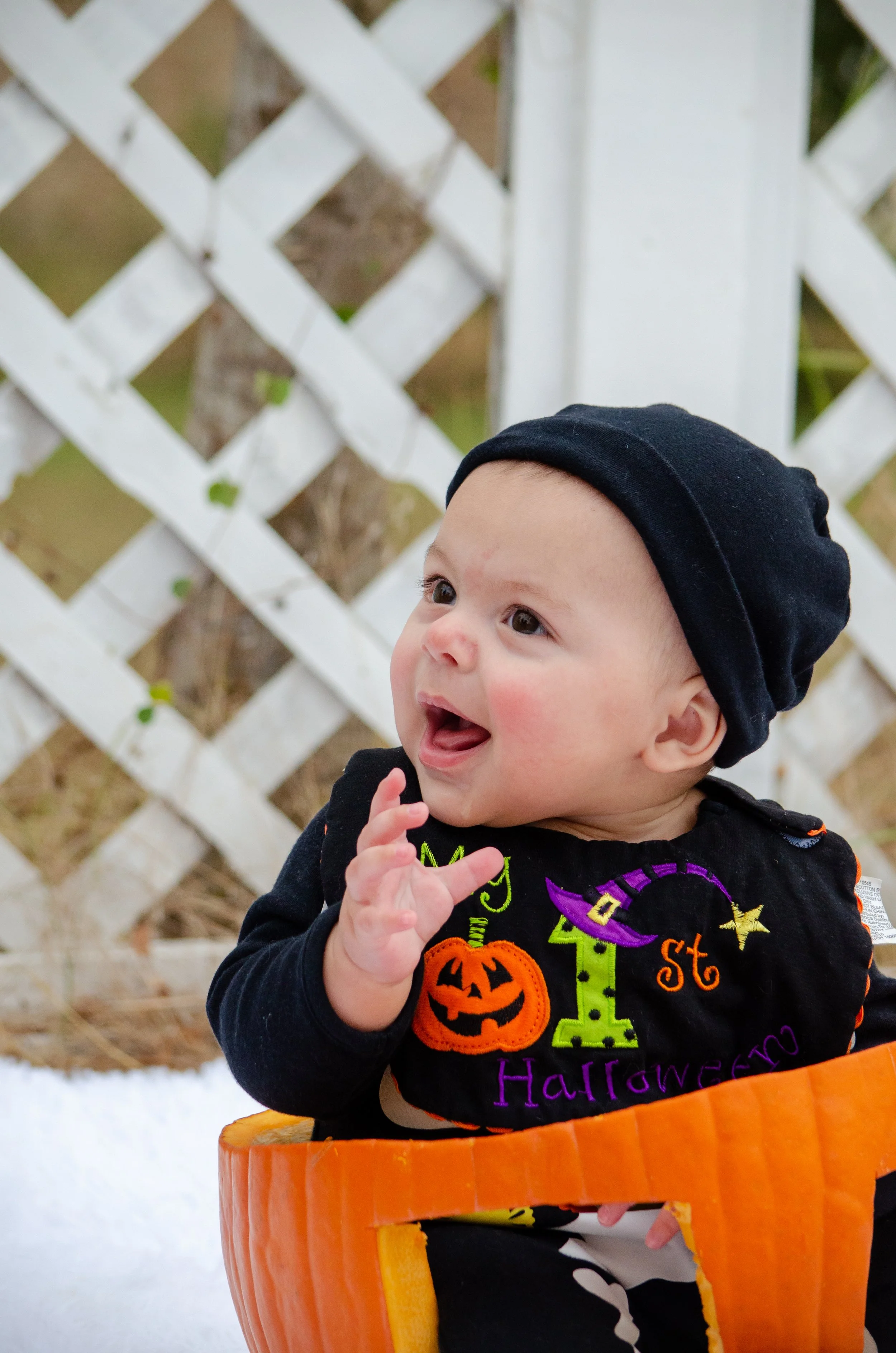 A smiling baby in a black Halloween shirt sitting in a pumpkin-shaped chair, wearing a black beanie, with a white lattice fence in the background.