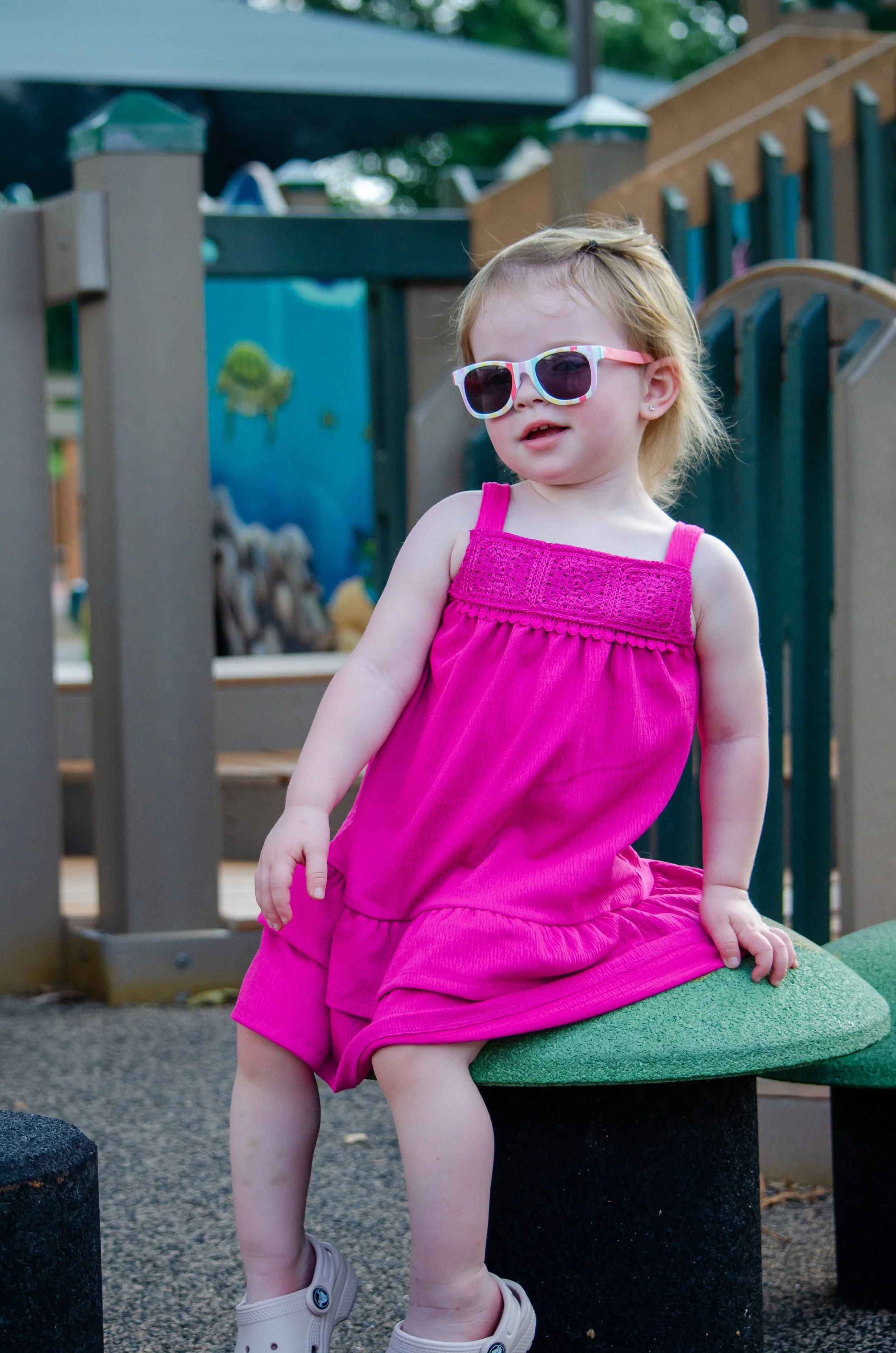 A young girl with blonde hair wearing sunglasses and a bright pink dress, sitting on a green and black playground structure during daytime.