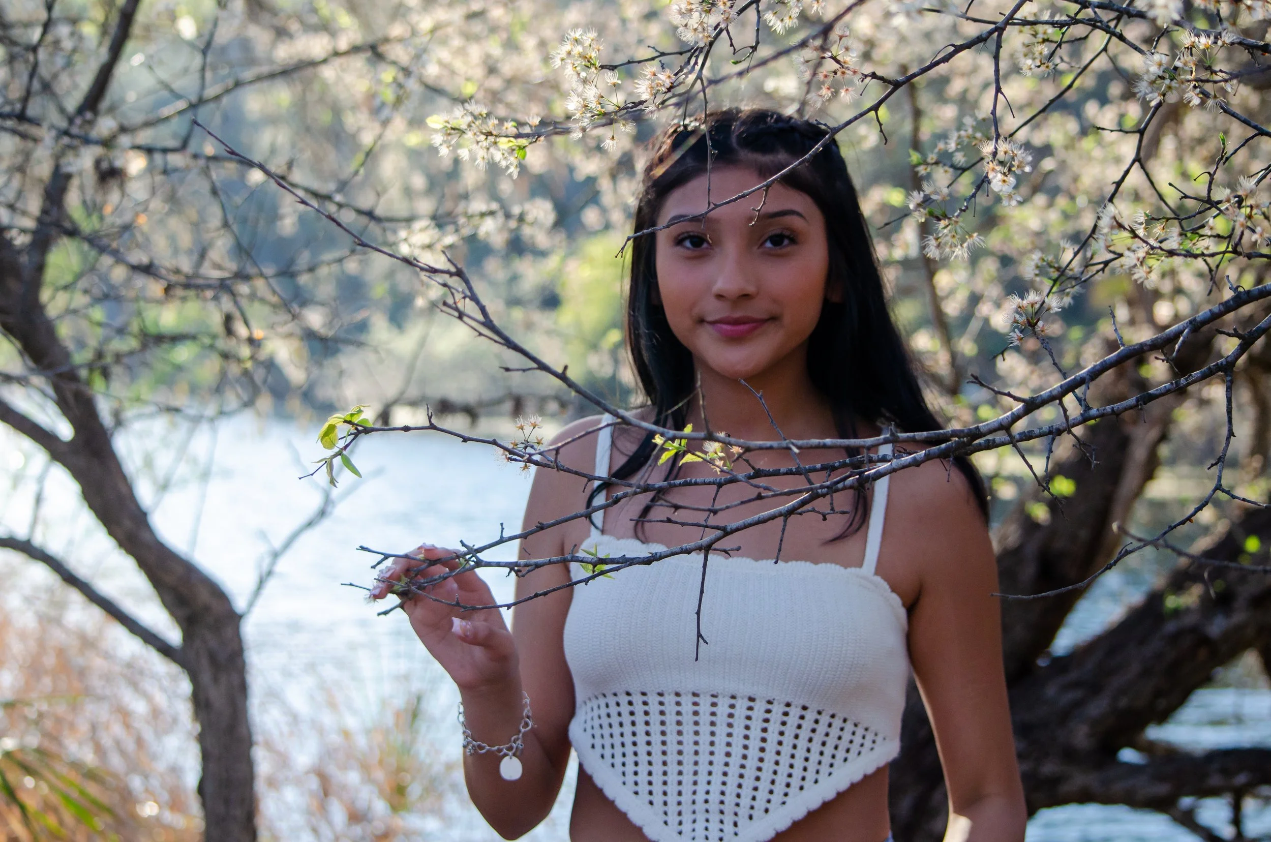 Young woman standing outdoors among blossoming trees by a body of water, holding branches and smiling at the camera.