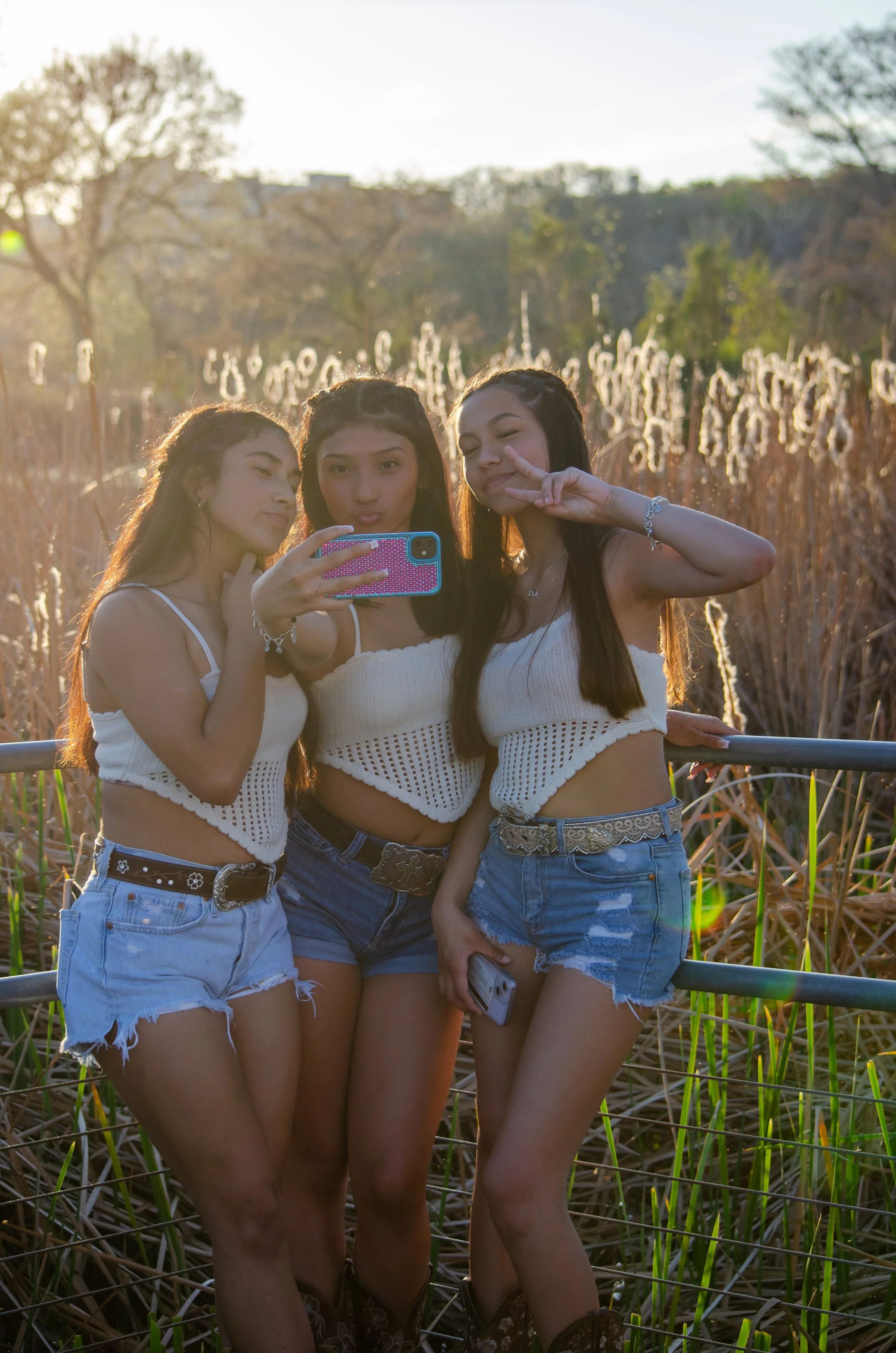 Three young women taking a selfie in a field during sunset, wearing white tops and denim shorts, making peace signs and posing for the camera.