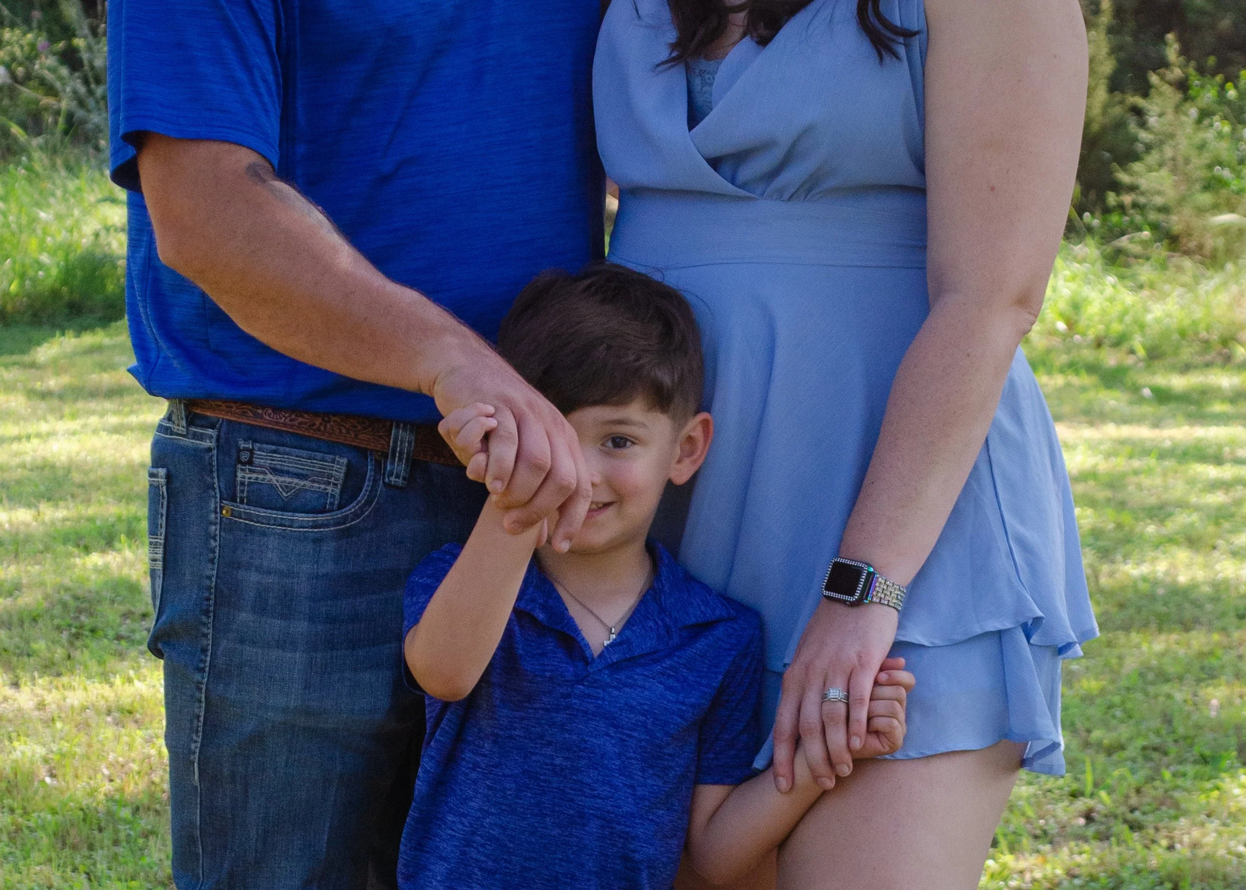 A young boy smiling while holding hands with a woman, with a man partially visible next to them, outdoors on a grassy area.