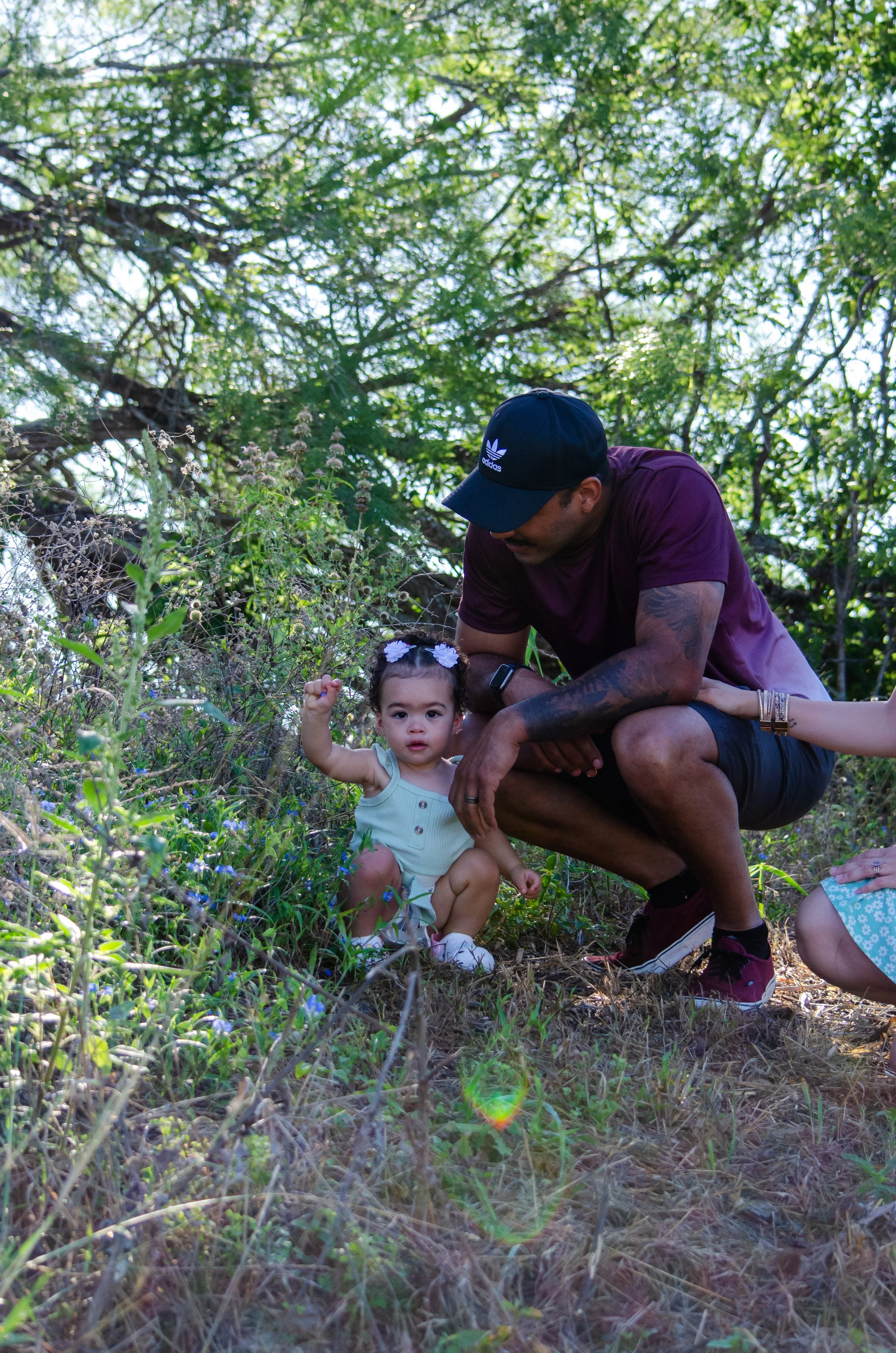A man crouching next to a young girl with dark curly hair and a flower headband, in a natural outdoor setting with green trees and plants. The girl is looking at the camera, raising her right hand, while the man is looking down at her.