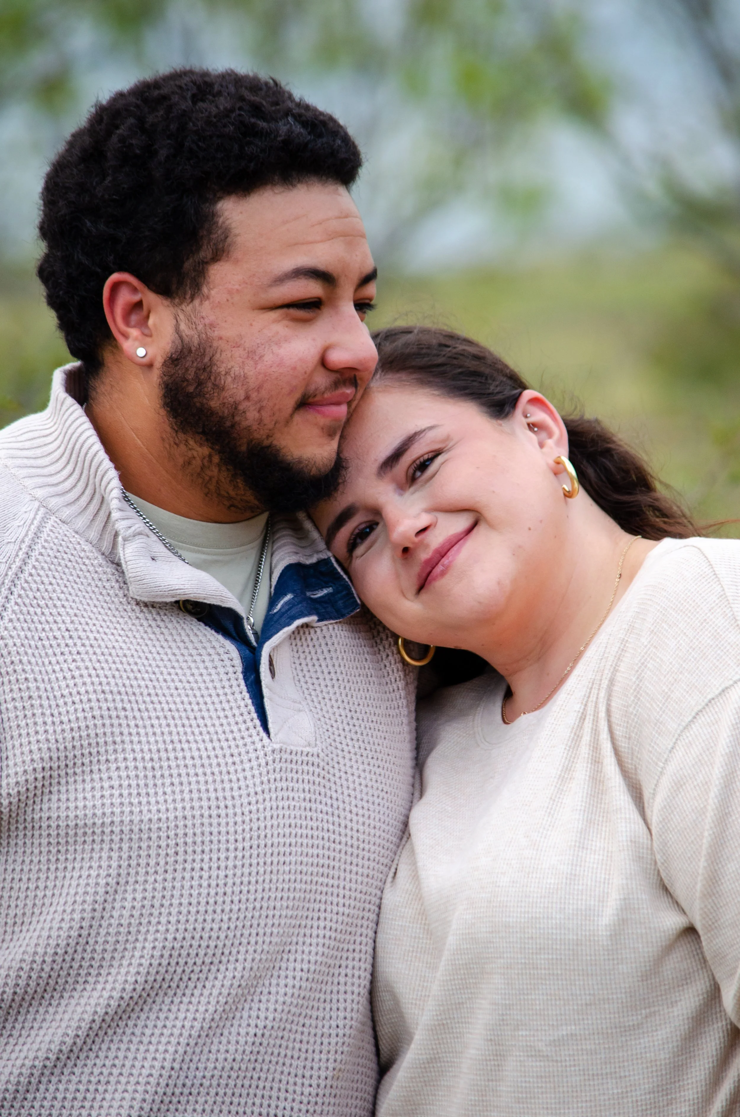 A young man and woman are close together outdoors, smiling and leaning their heads against each other.
