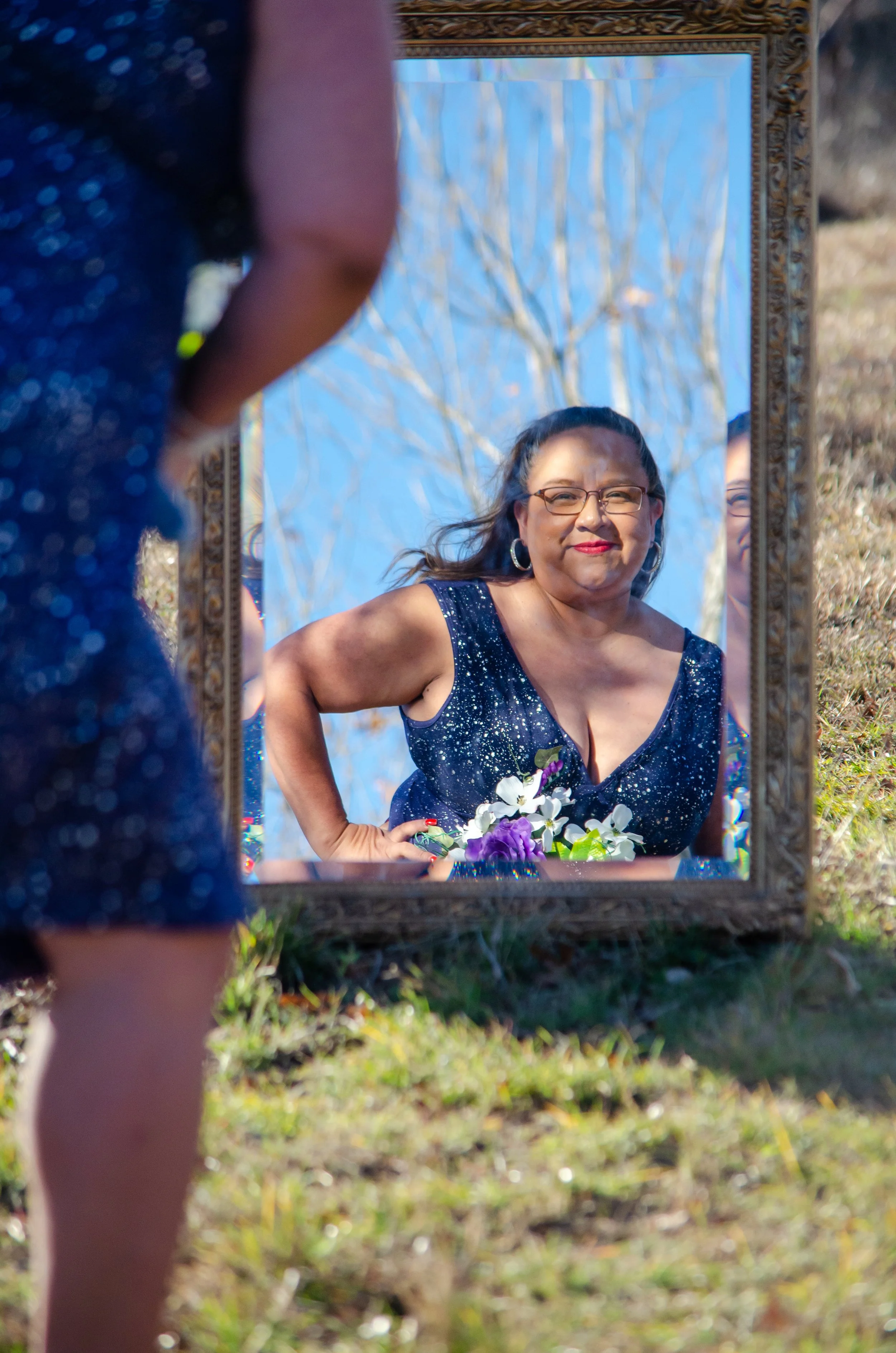 A woman in a blue sparkly dress looking at her reflection in an outdoor mirror, holding a bouquet of flowers, with bare trees and blue sky in the background.
