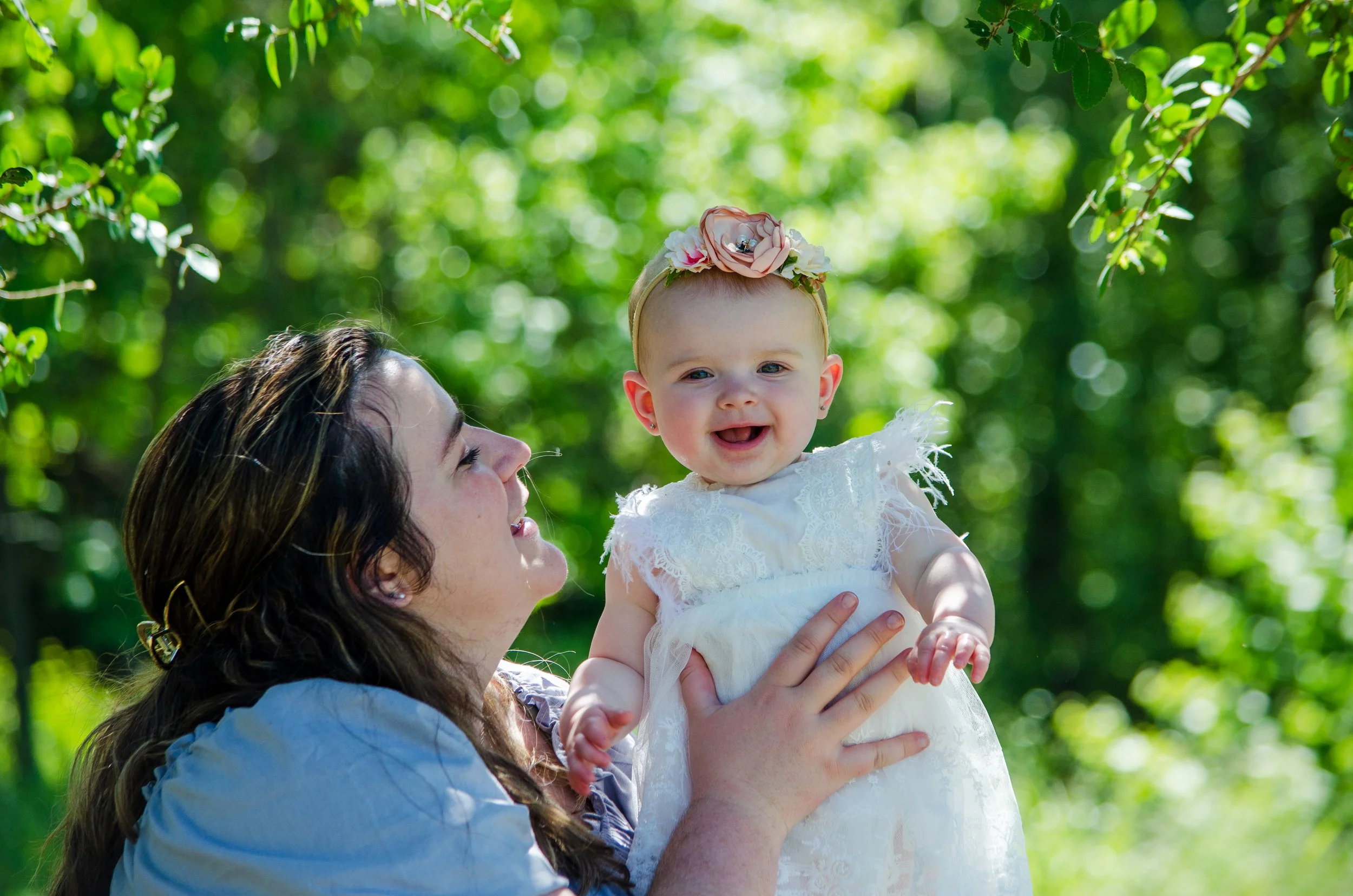 A woman lifting a smiling baby girl outdoors among green trees, wearing a white lace dress and pink floral headband.