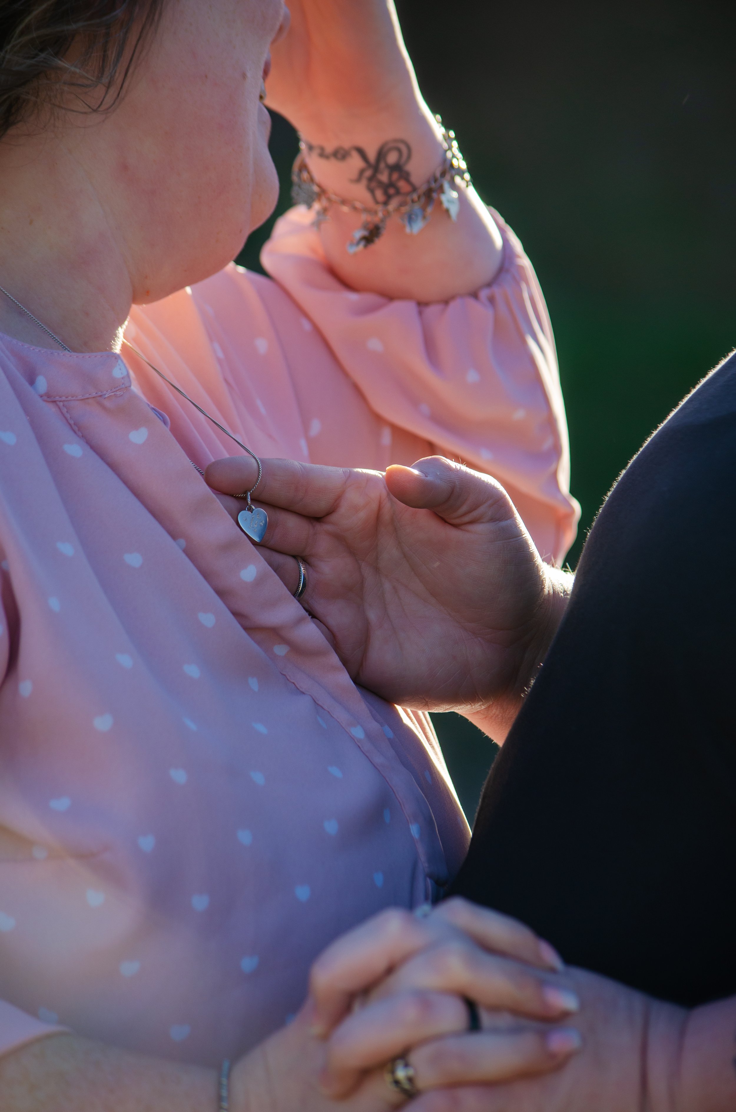 A close-up of a couple dancing outdoors, with one person's hand on the other's chest and the other person holding the other's hand. The woman is wearing a pink polka-dot dress and jewelry, and the scene is lit by natural sunlight.